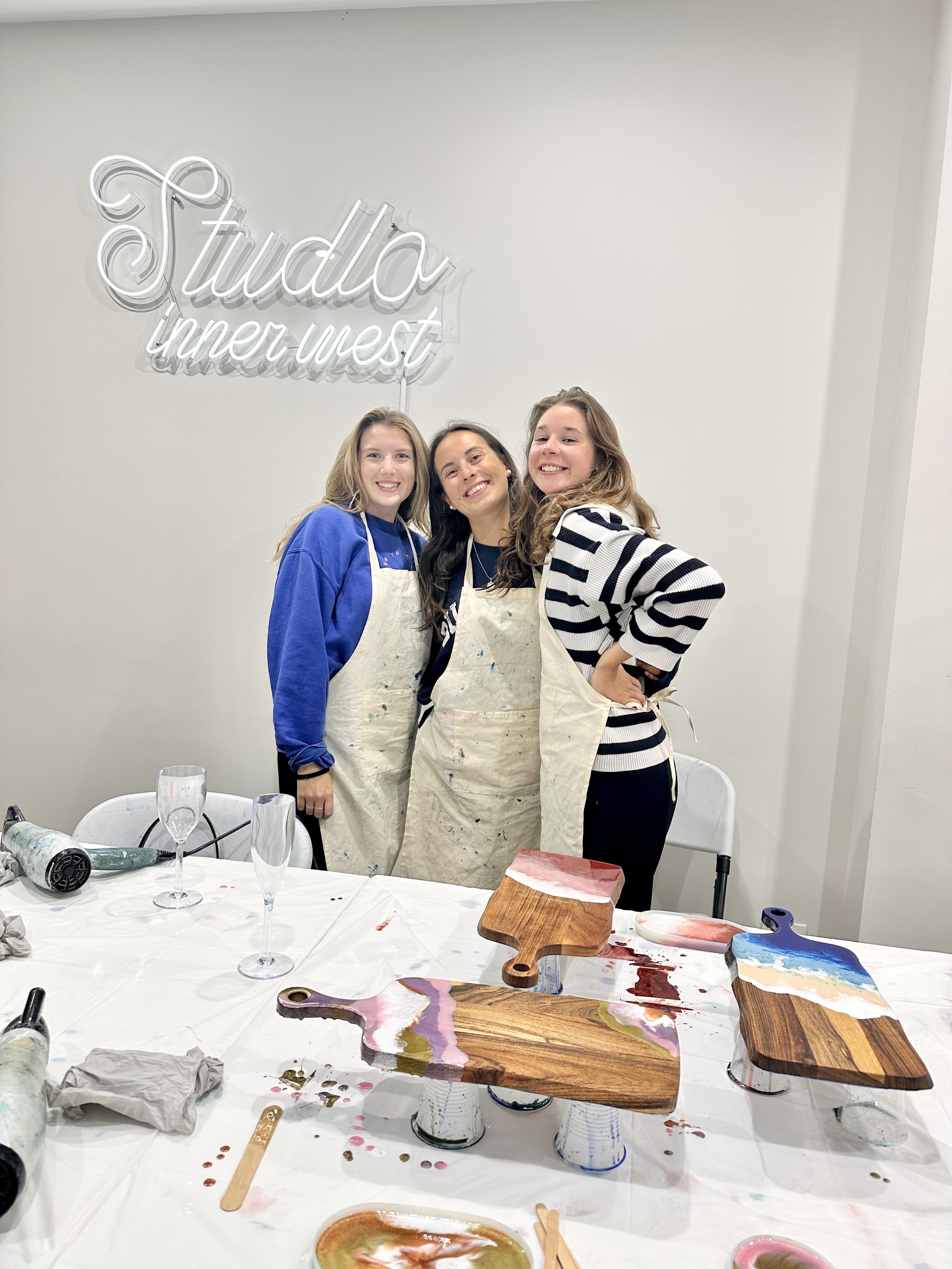 Three people wearing aprons stand smiling in front of a wall sign that says "Studio inner west." The table in front of them features art supplies and decorated wooden cutting boards.