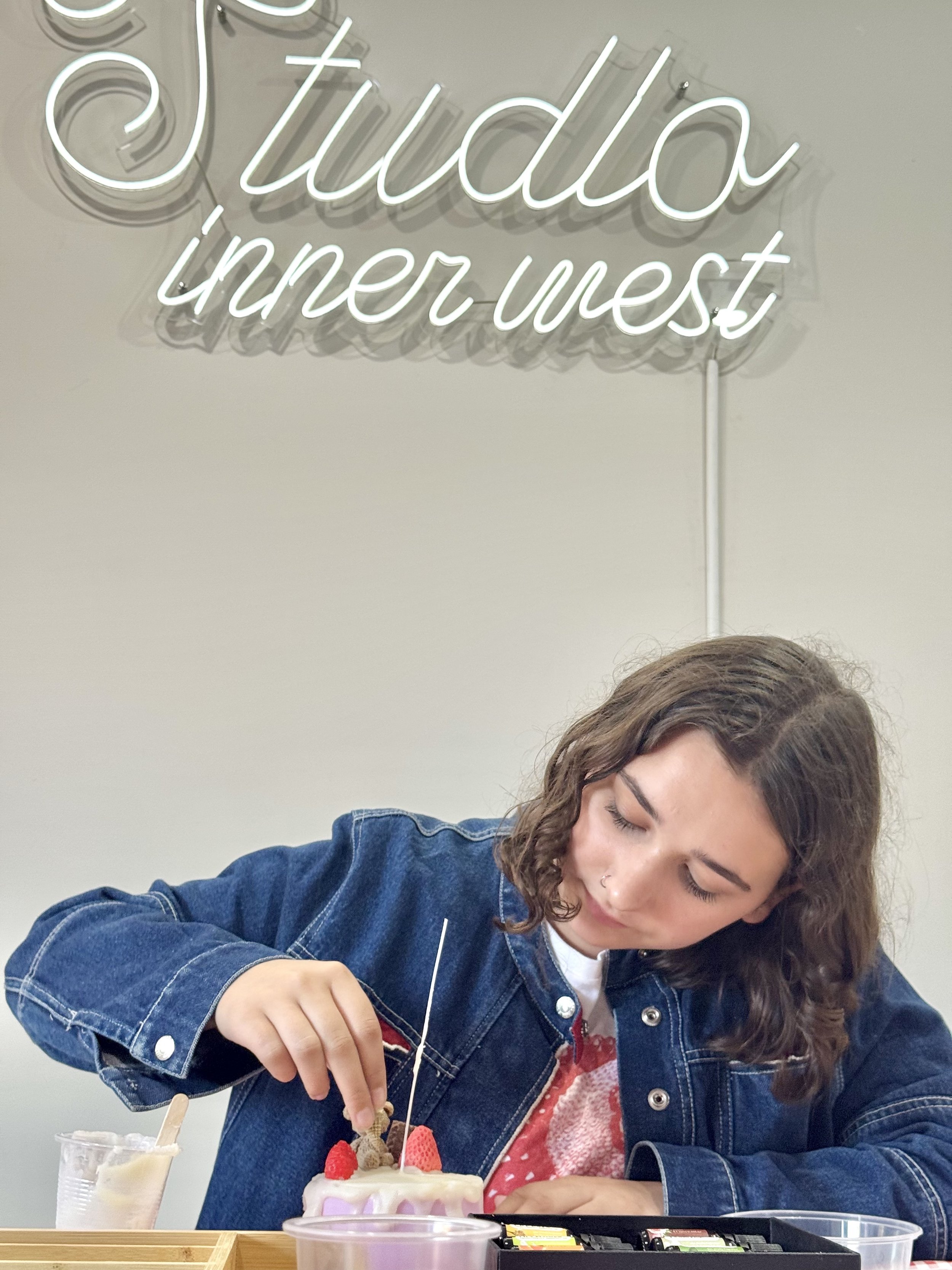 Person in a denim jacket decorating a purple cake with strawberries and other toppings, under a neon sign that reads "Studio inner west."