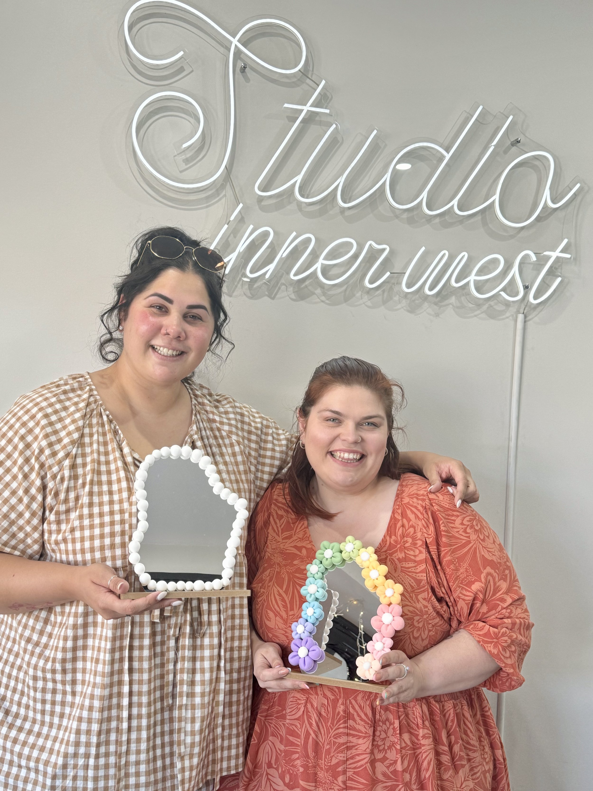 Two people smiling and holding decorative mirrors, one adorned with white beads and the other with colorful flowers, standing in front of a sign reading 'Studio Inner West.'