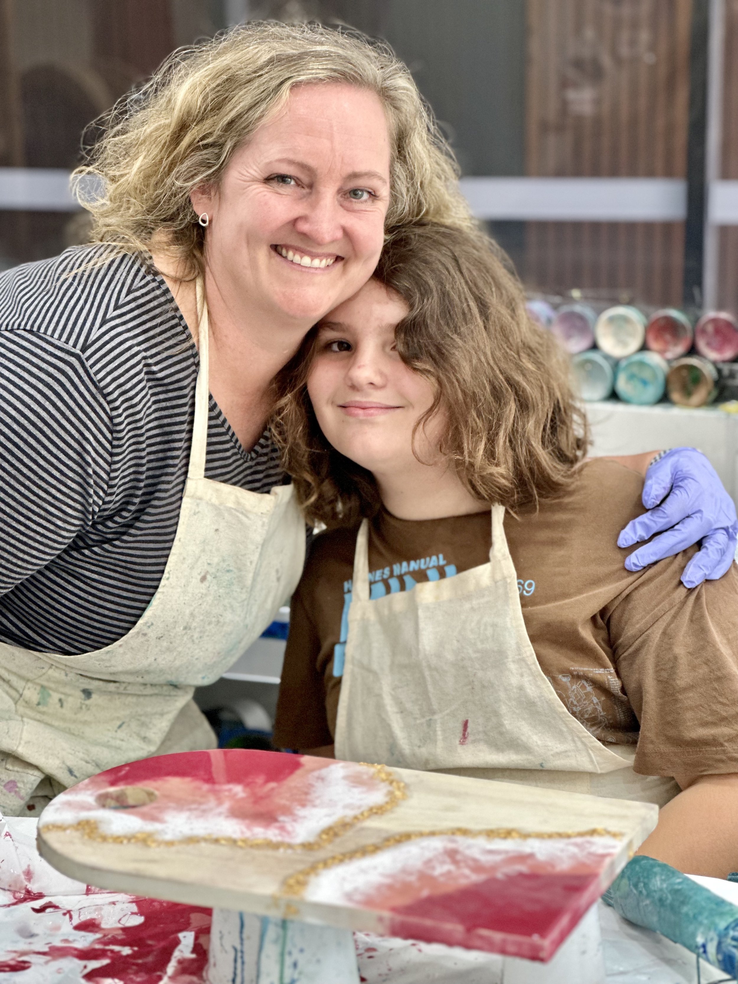 Two people wearing aprons, standing close together and smiling in an art studio. They are near a table with paint supplies, including a canvas with red and white colors and a gold line. One person is wearing a striped shirt, the other a brown shirt.