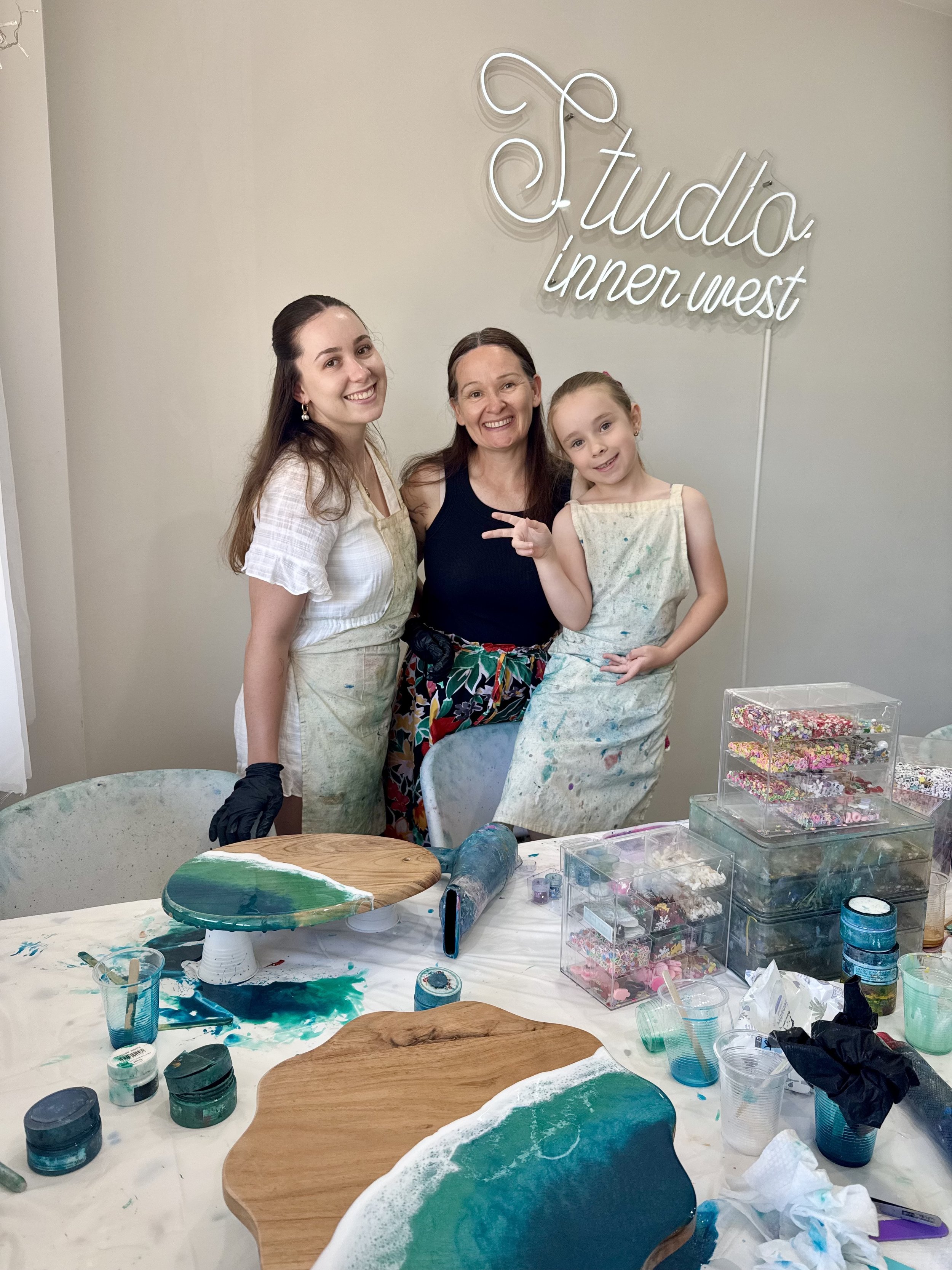 Three women standing next to a craft table with art supplies and decorated wooden boards, in a room with a "Studio inner west" sign on the wall.