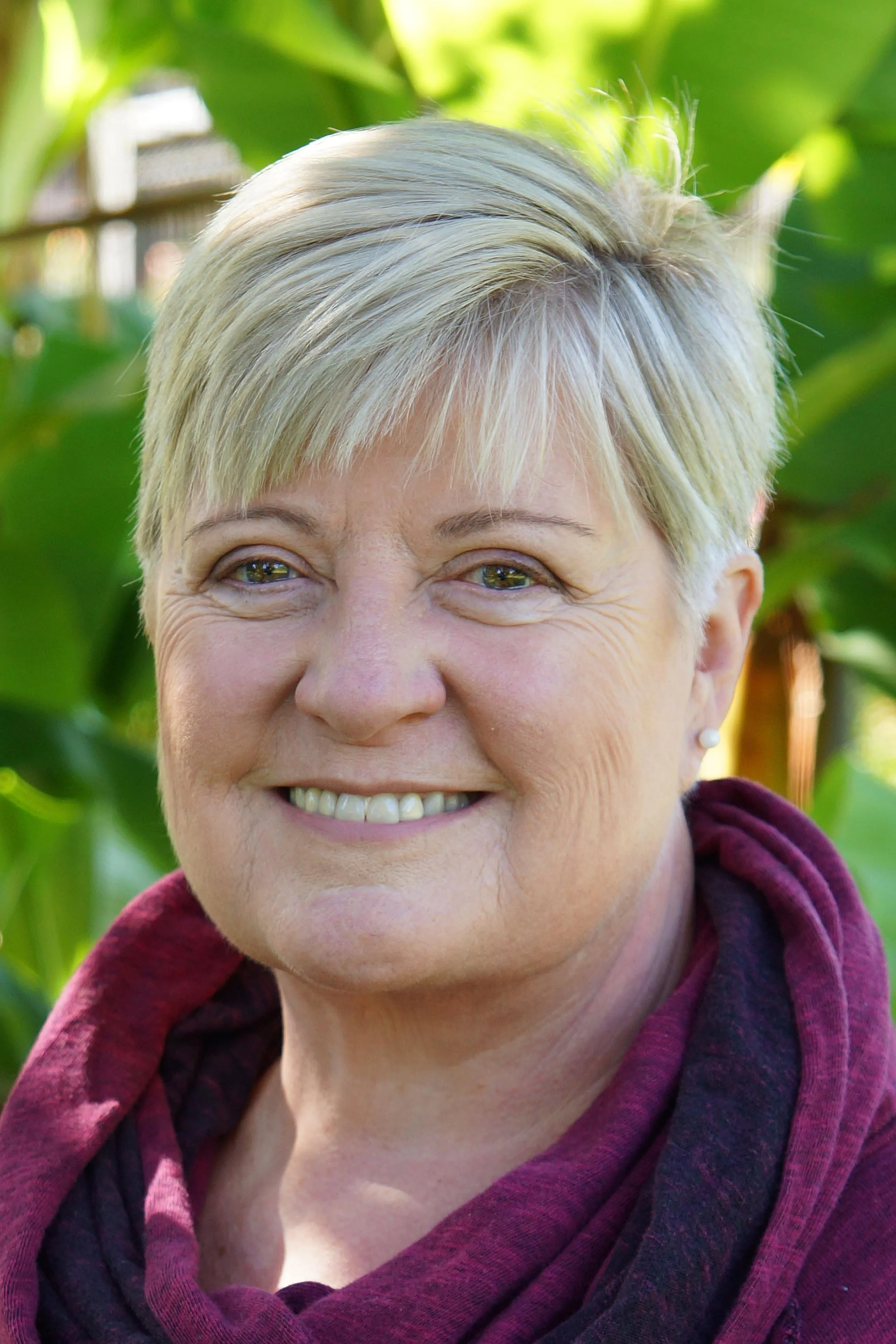 A smiling woman with short blonde hair, wearing a maroon scarf, outdoors with green foliage in the background.