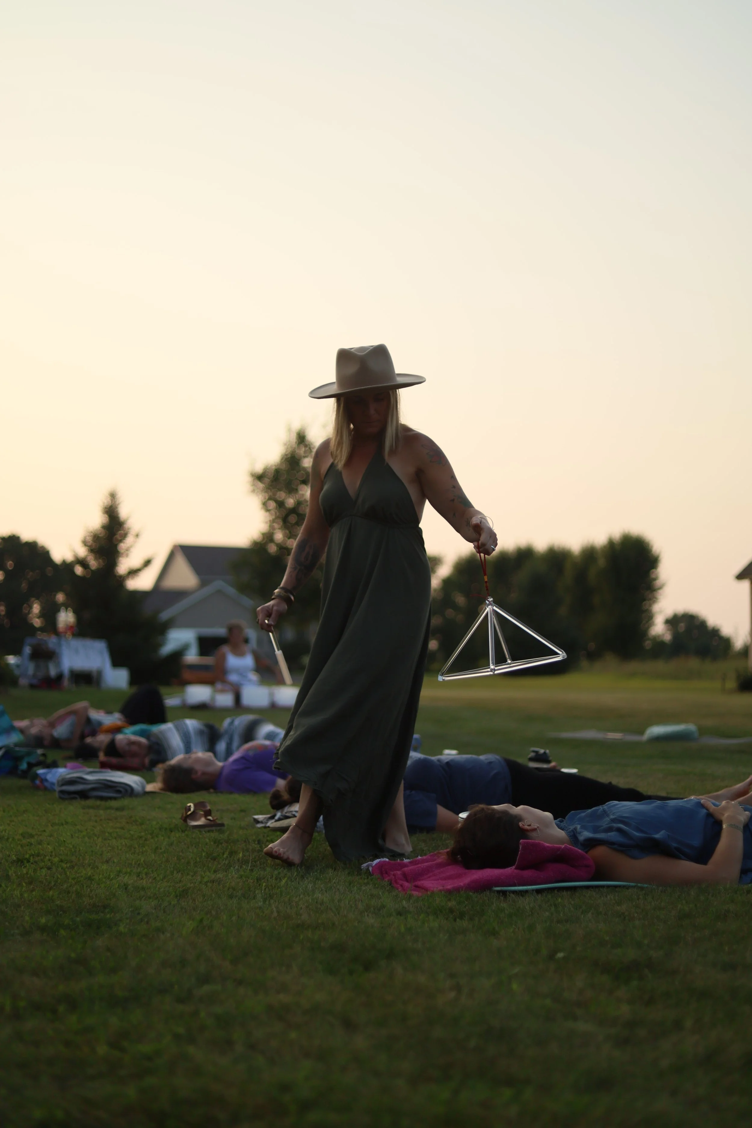 A woman with blonde hair, wearing a wide-brimmed hat and a long dark dress, is walking across a grassy field hosting yoga class. She appears to be holding a metallic geometric kite. People are lying on mats and blankets, practicing yoga, during sunset or early evening, with houses and trees in the background.