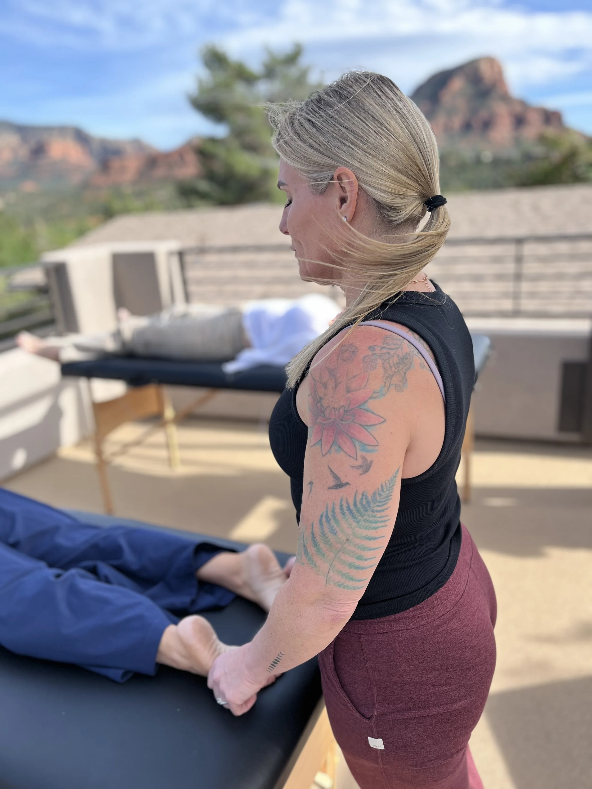 A woman with blonde hair tied back, tattoos on her arm, wearing a black tank top and maroon leggings, holds the hands of a person lying on a table. The person lying down is out of focus, but appears to be receiving some form of treatment or therapy. The background features an outdoor setting with a wooden fence, landscape, and rock formations in the distance under a partly cloudy sky.