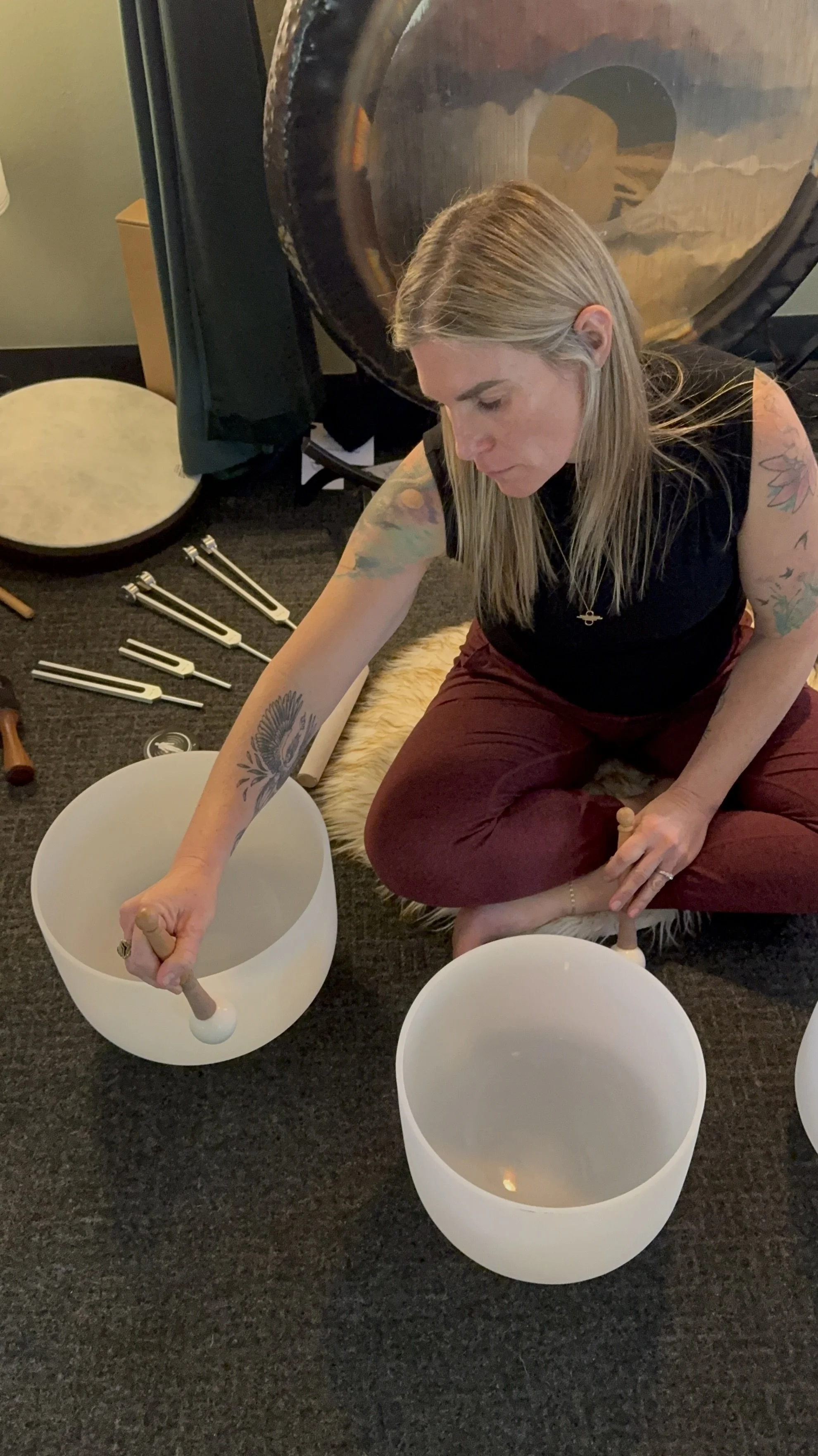 A woman with long blonde hair and tattoos on her arms is sitting on the floor, playing crystal singing bowls with a mallet. She is surrounded by musical instruments and a large gong in the background.
