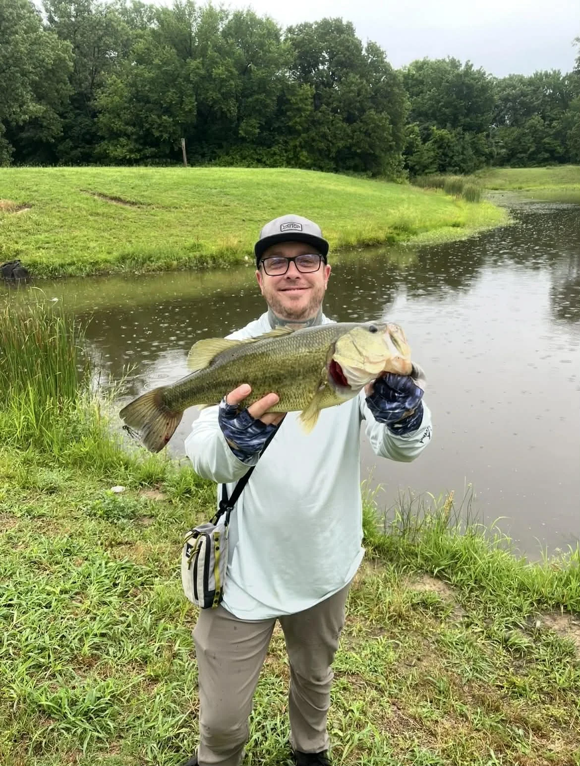 A man standing by a pond holding a large fish he caught, with trees and grassy landscape in the background.