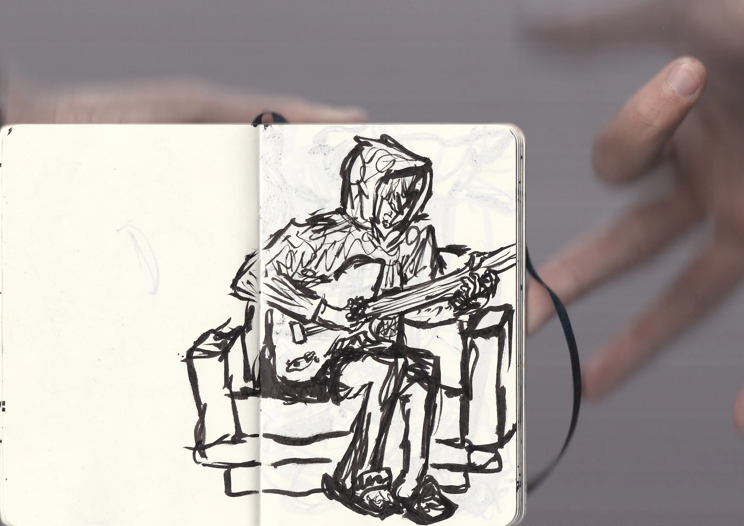 A person's hand holding an open sketchbook with a black ink drawing of a person sitting at a table, wearing a hooded jacket, with a plate and other items on the table.