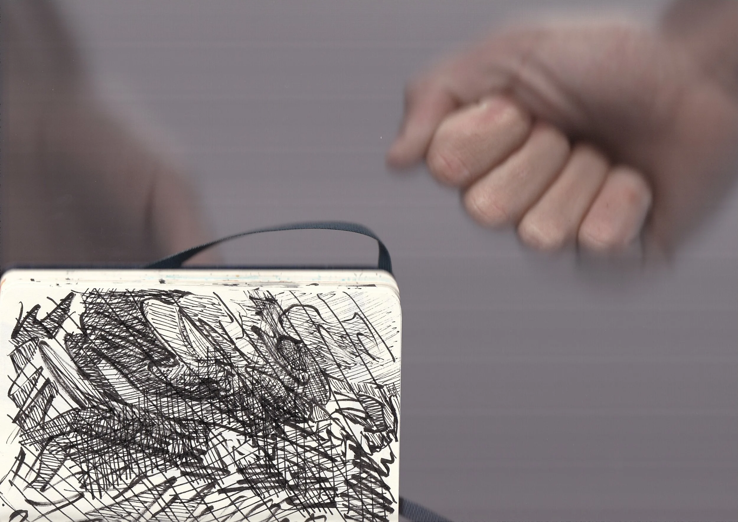 Close-up of a hand holding a white card with black scribbles, with another hand in the background, and part of an electronic device at the bottom.