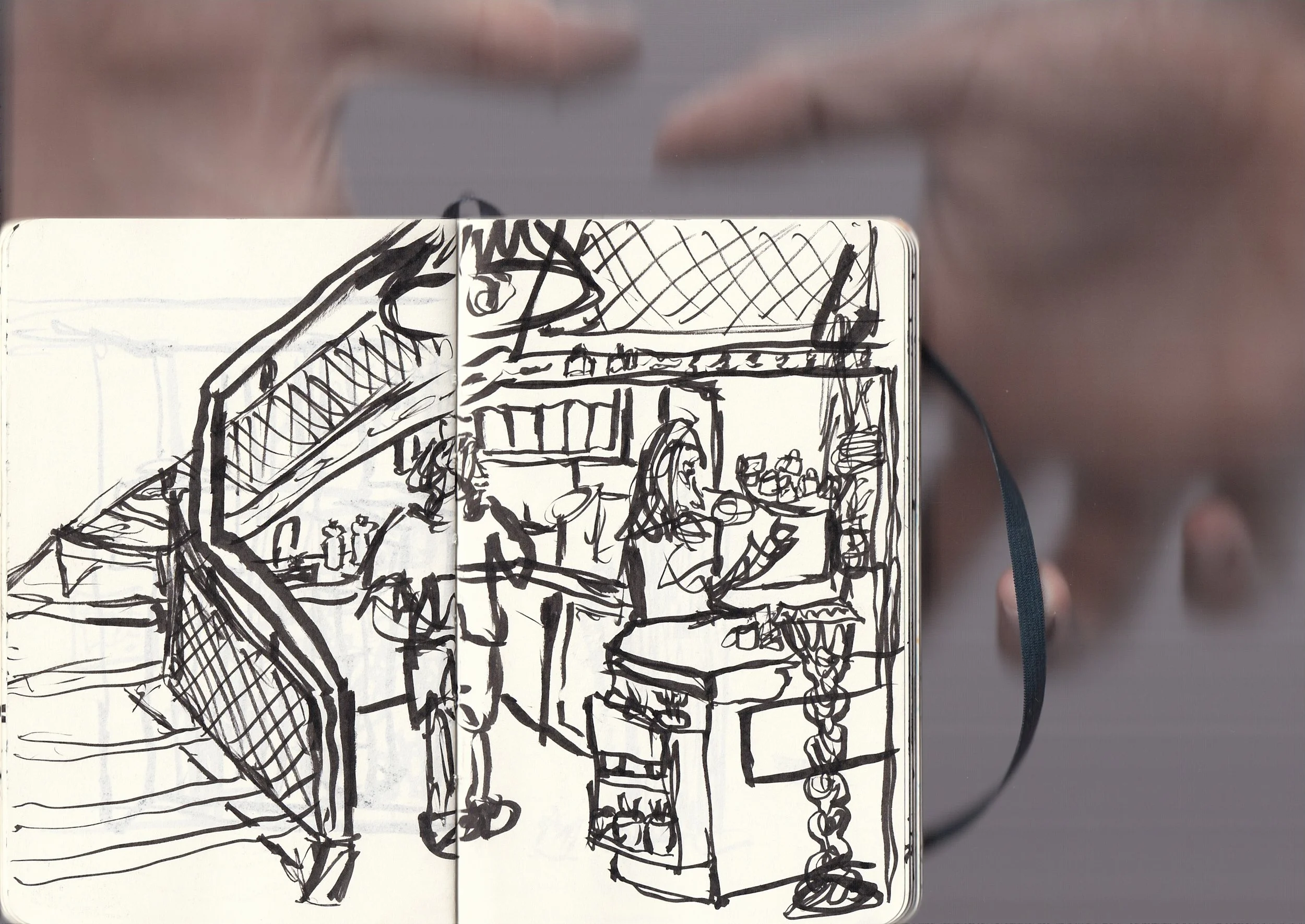A sketch of an outdoor scene with tables and chairs, some people, and a large structure, drawn with black ink on an open notebook.