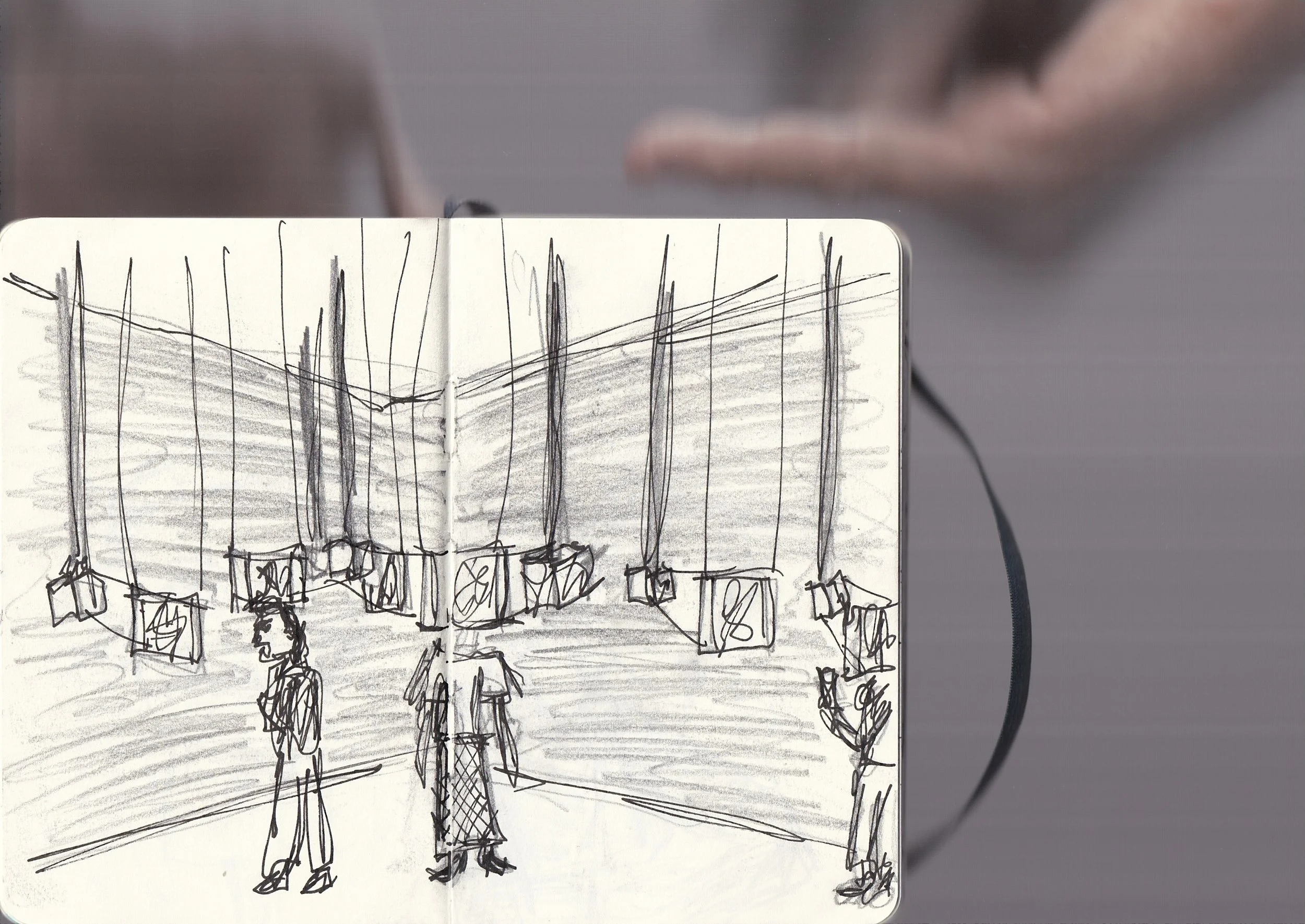 A sketched drawing of people walking in front of a large array of hanging lights in a public space. The scene is depicted on a piece of paper being held up or viewed from in front of a person.