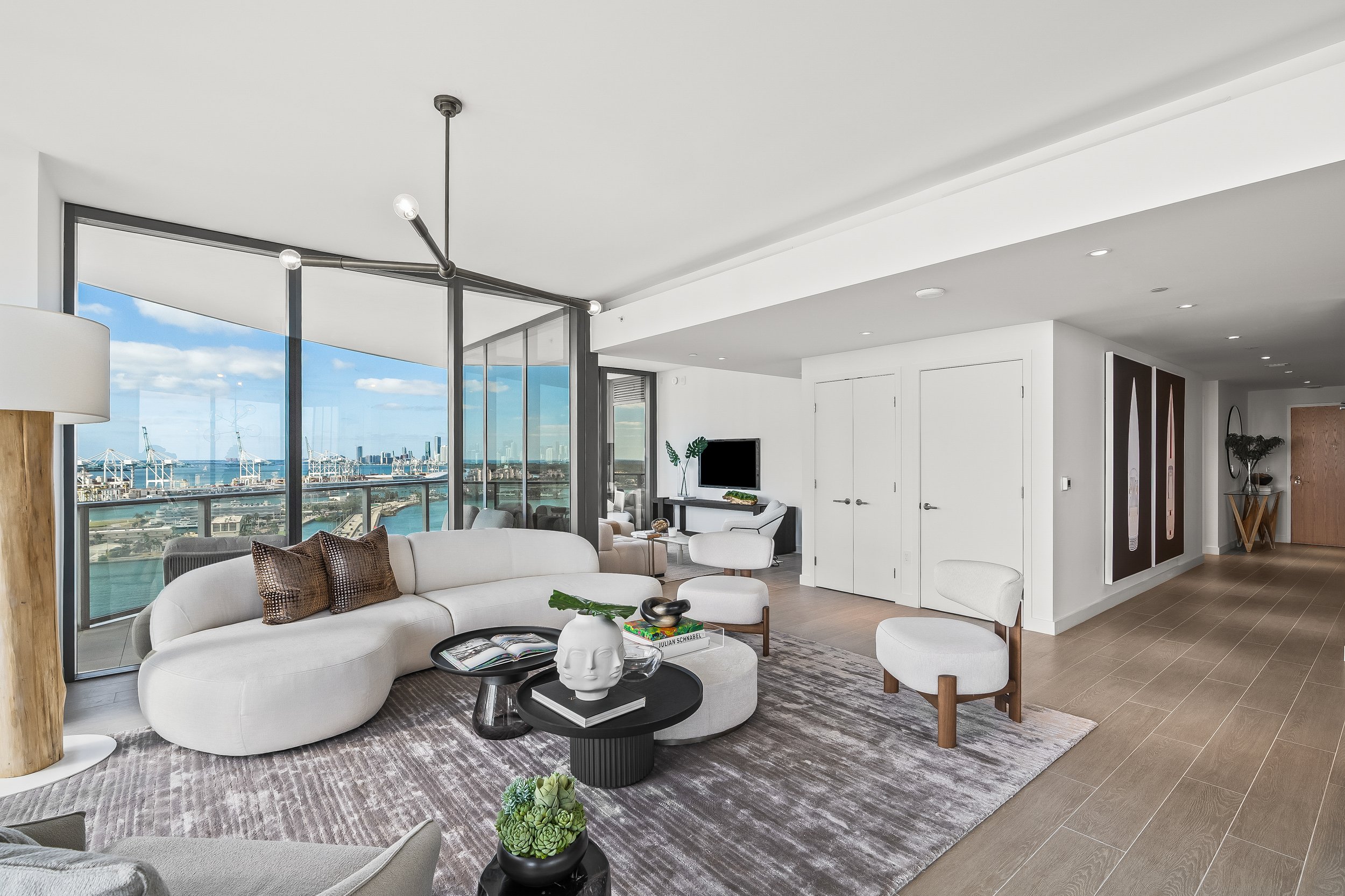 Modern living room with panoramic view, white sectional sofa, circular coffee tables, decorative pillows, abstract art, and large windows overlooking city and water.