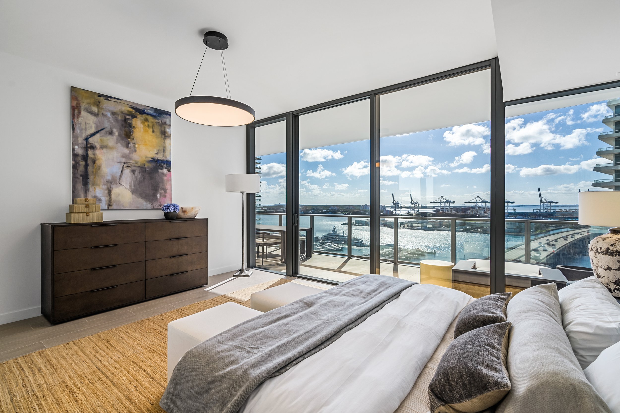 Modern bedroom with large windows overlooking a harbor view, featuring a bed with gray and white bedding, a textured rug, a dark wooden dresser, abstract wall art, and ceiling lighting.