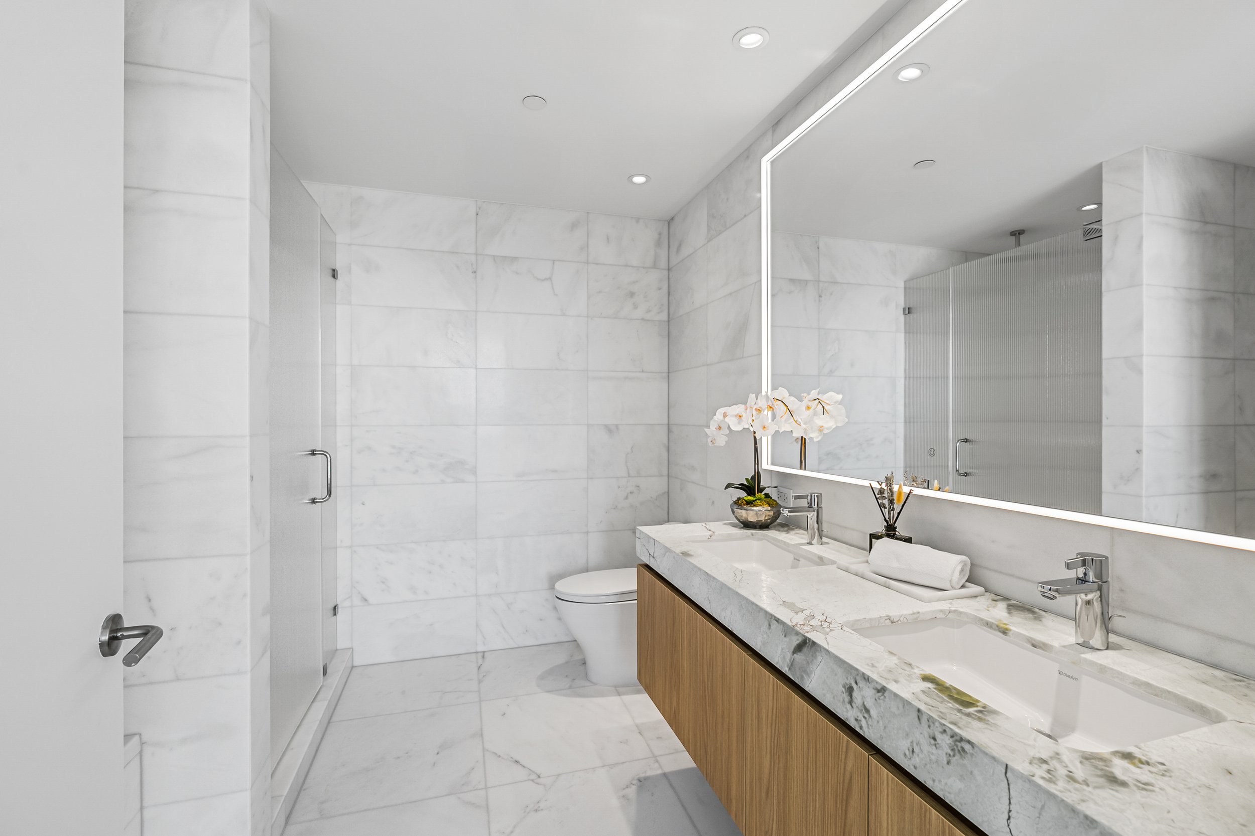 Modern bathroom with marble tiles, large mirror, double sink vanity, white toilet, and a glass-enclosed shower.