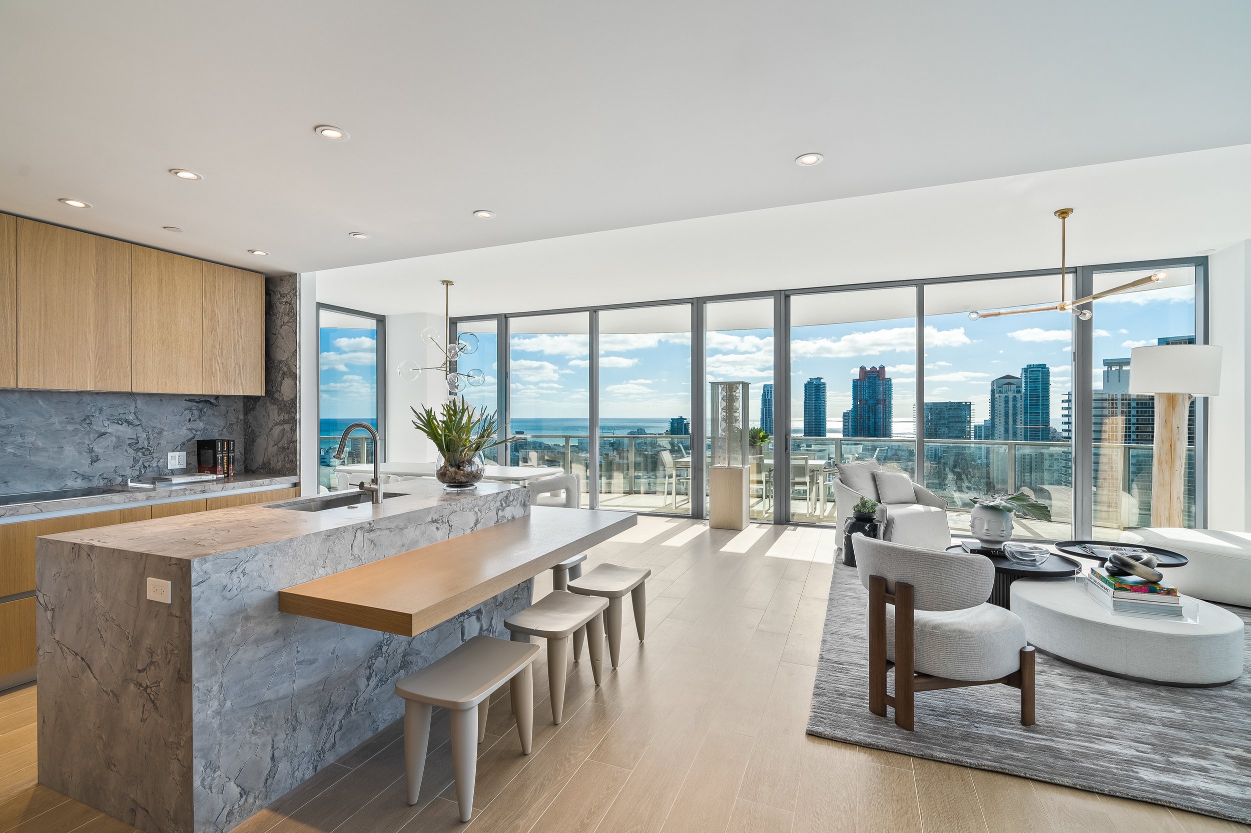 Modern open-plan living space with kitchen and large windows, featuring light wood and marble finishes, contemporary furniture, and cityscape view.