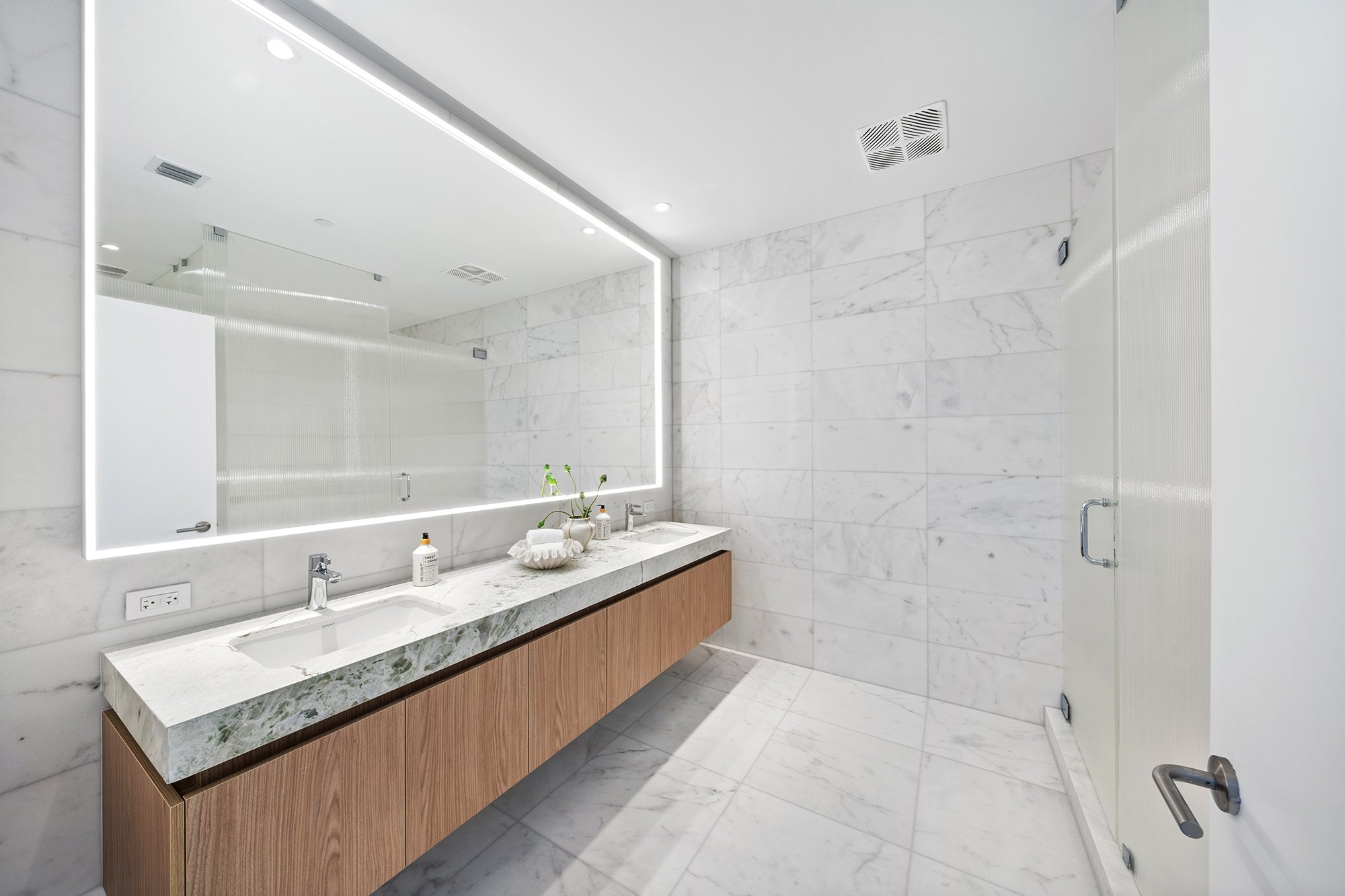 Modern bathroom with a large illuminated mirror, marble countertops, and wooden cabinetry, featuring a sleek glass shower door and marble tile walls.
