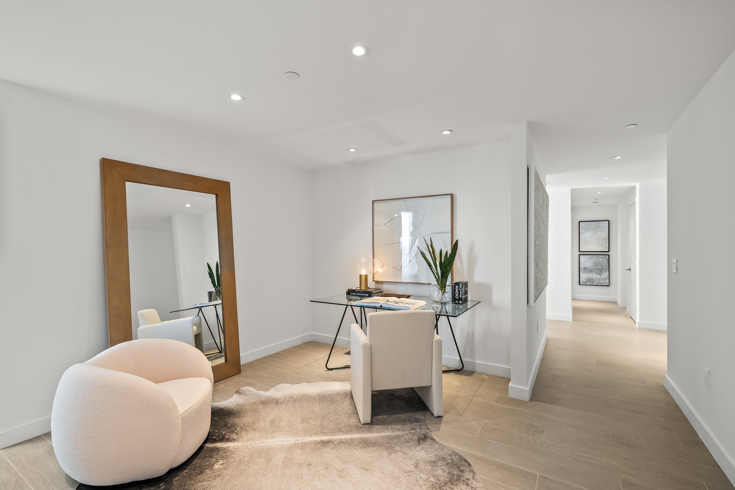 Modern home office with a large mirror, white armchair, glass desk, decorative plant, and wall art. Light wood flooring and bright neutral decor.