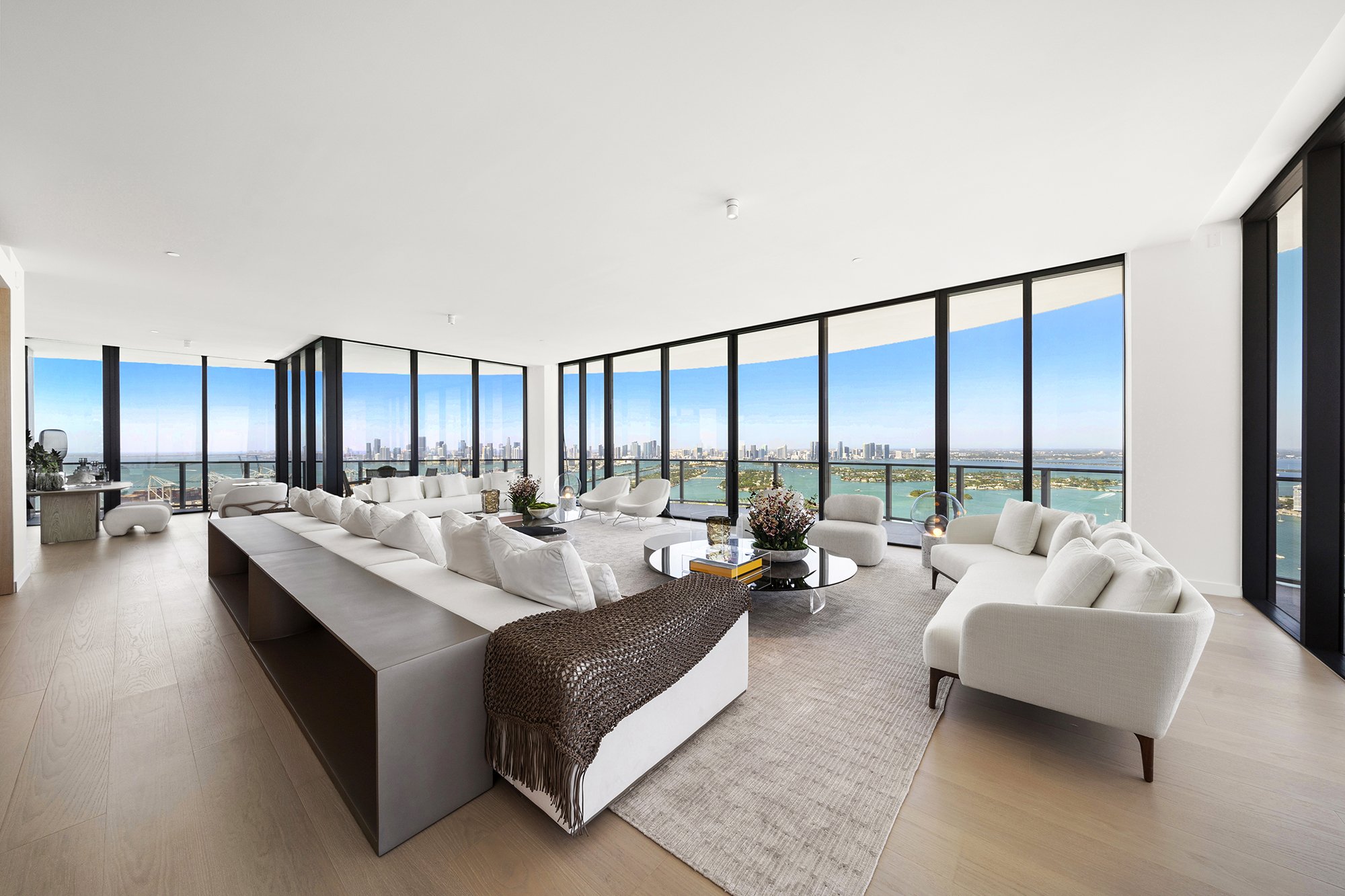 Modern living room with large windows, white sofas, armchairs, glass coffee tables, and a city and water view.