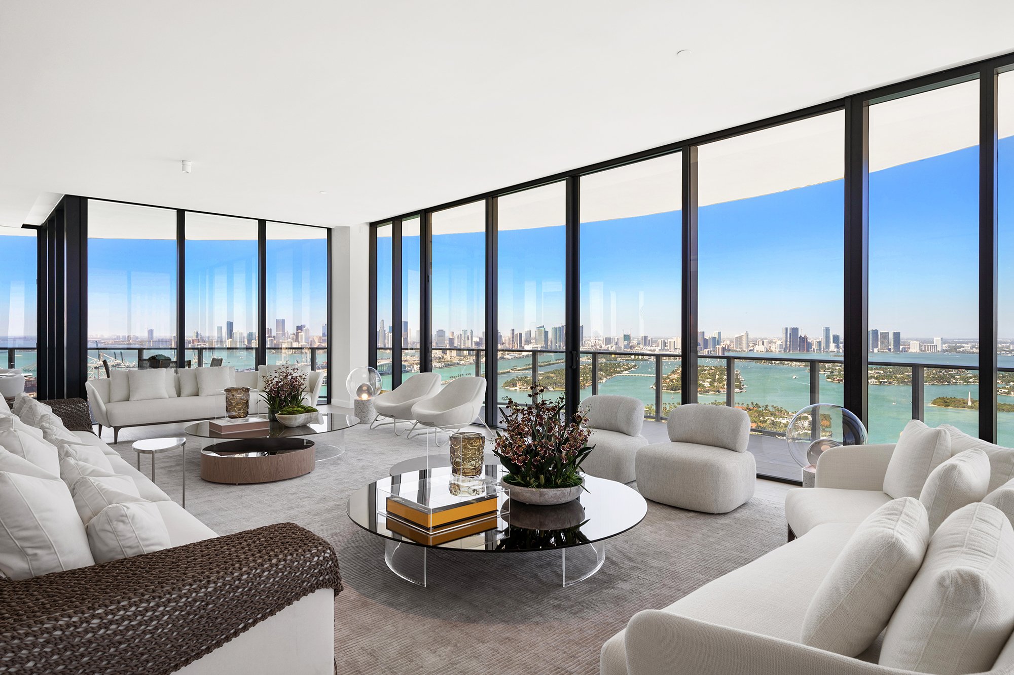 Modern living room with large windows overlooking a city skyline and water, featuring white sofas, armchairs, and round coffee tables with plants.
