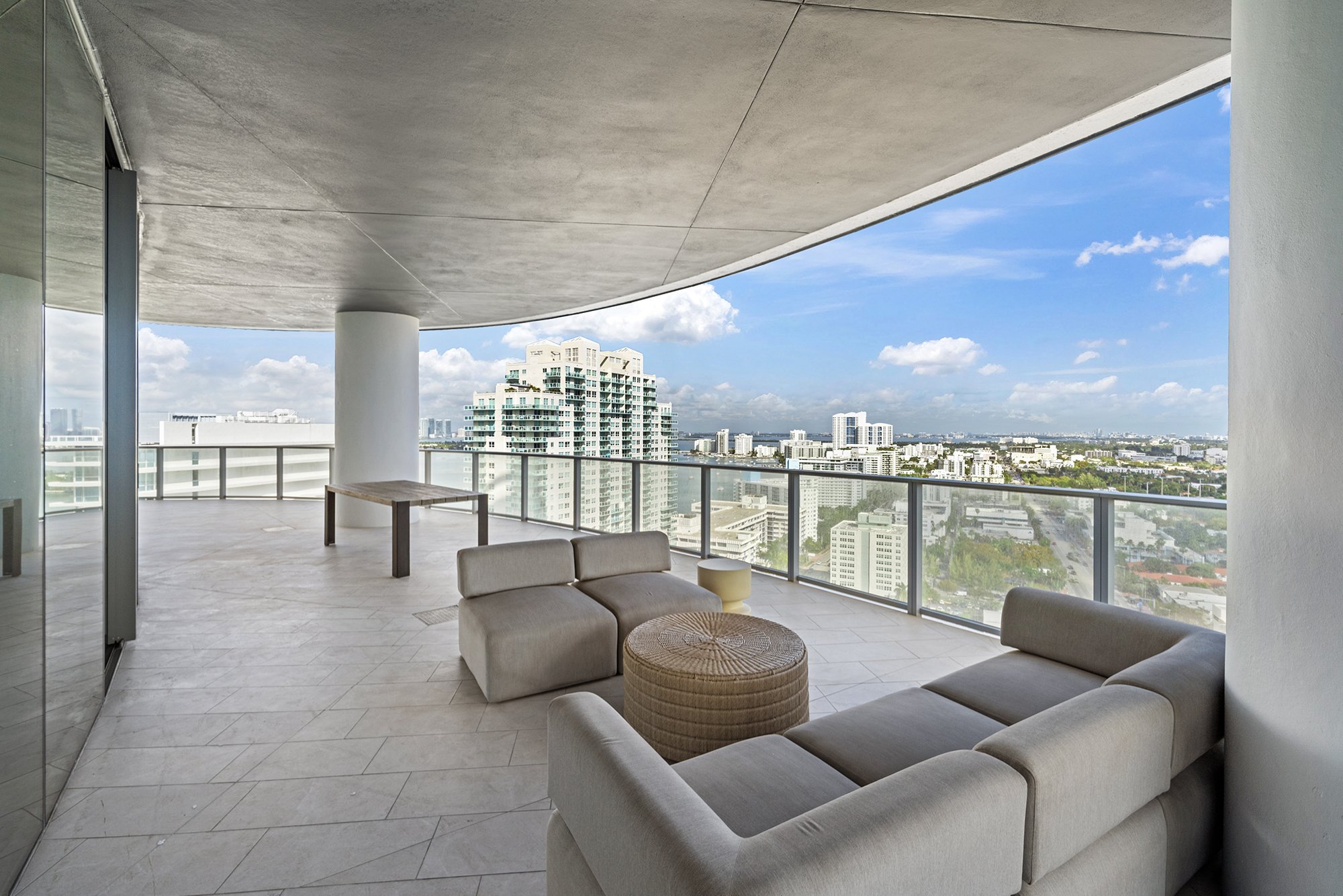 Modern apartment balcony with furniture, glass railing, and city skyline view under a clear blue sky.