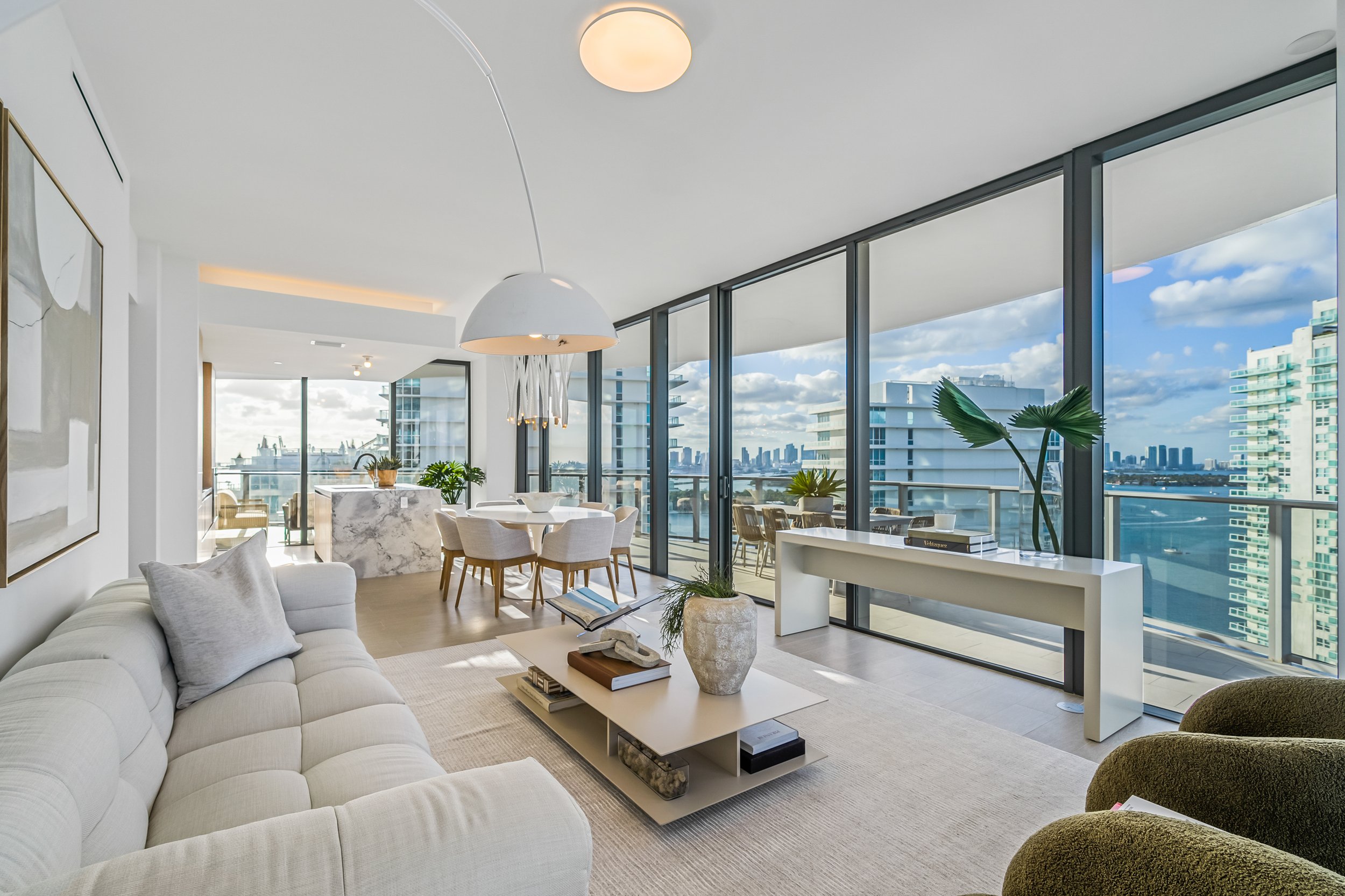 Modern living room with a beige sofa, glass walls, and a view of a city skyline over water. Features a white coffee table with decorative items and a dining area with a marble table and chairs. Contemporary ceiling light and green plants accent the s