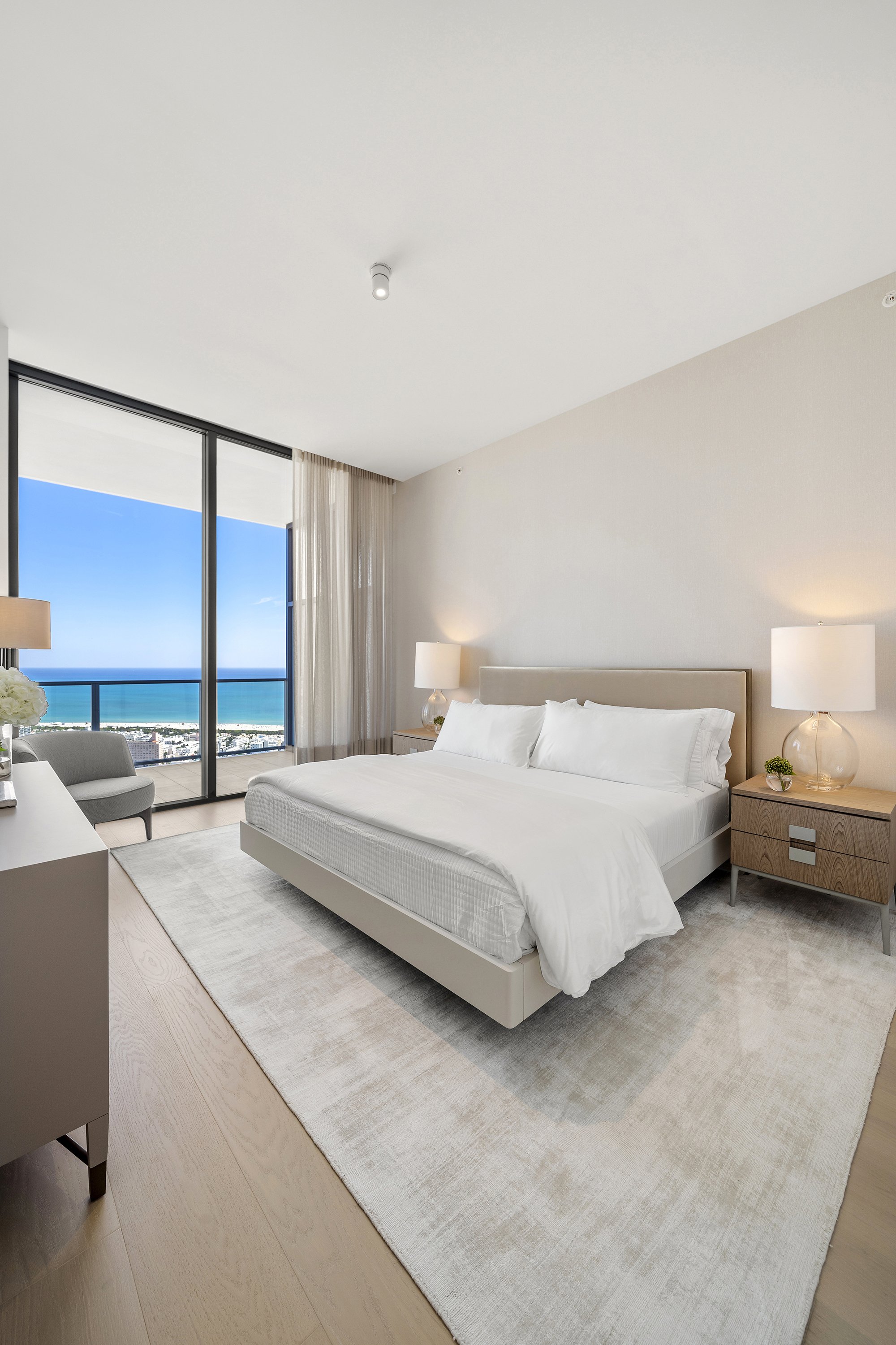 Modern bedroom with a large bed, bedside tables, lamps, and a panoramic ocean view through floor-to-ceiling windows.