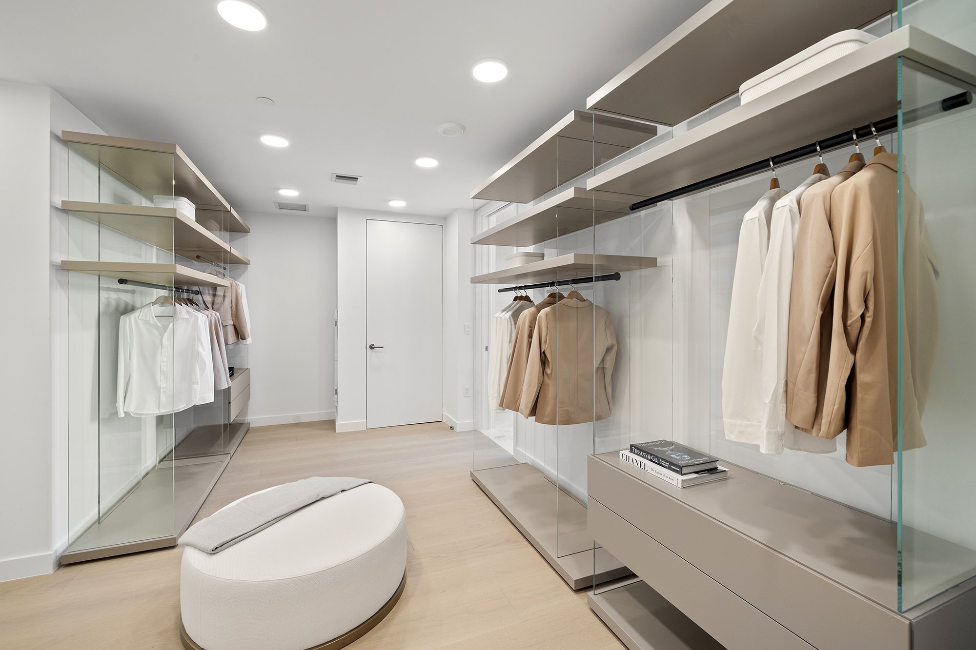 Modern walk-in closet with beige and white clothing on hangers, open shelving, a round ottoman, and a minimalist design.