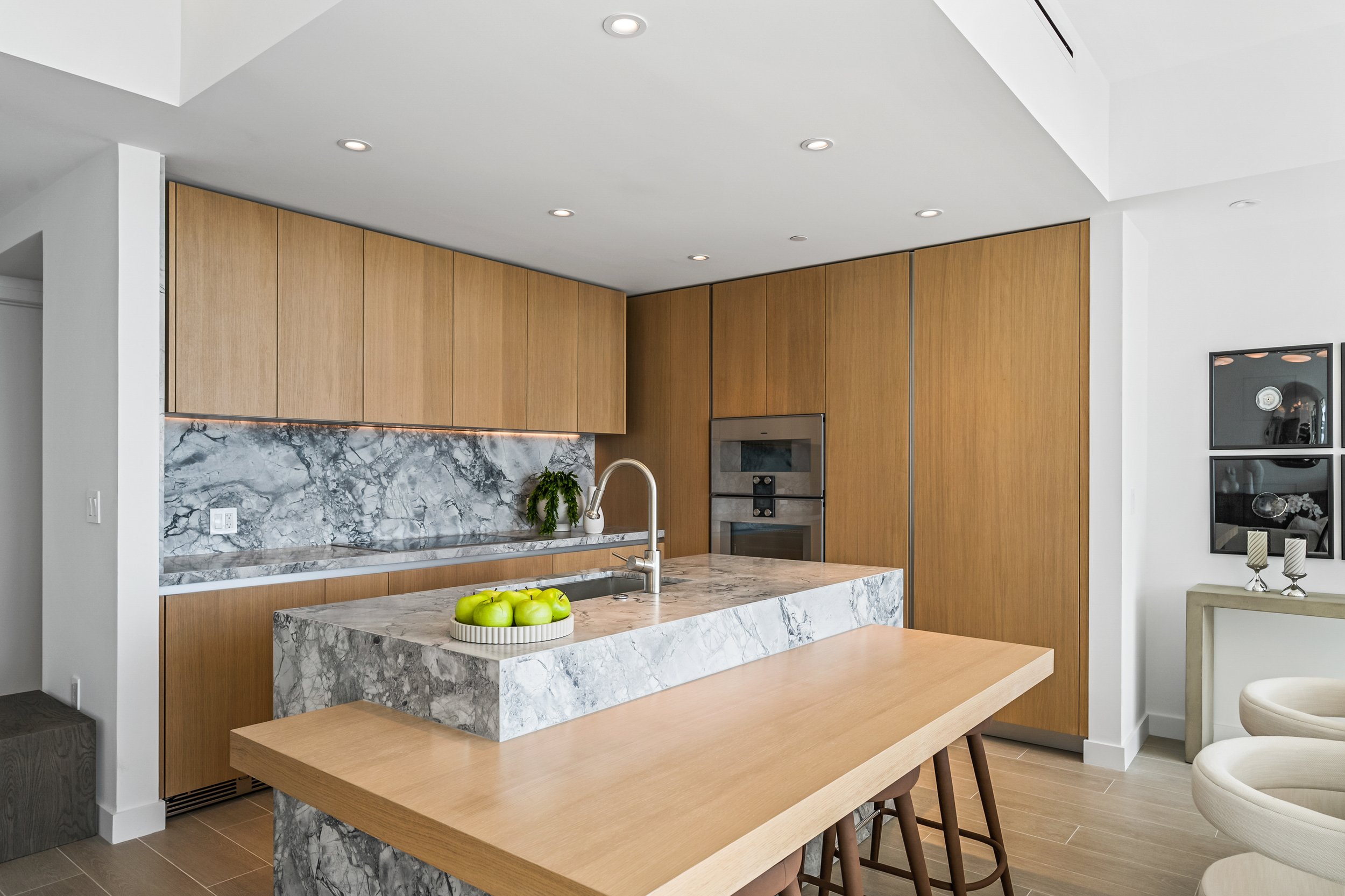 Modern kitchen with marble countertops, wooden cabinets, bar stools, and a stainless steel faucet. A bowl of green apples is on the counter.