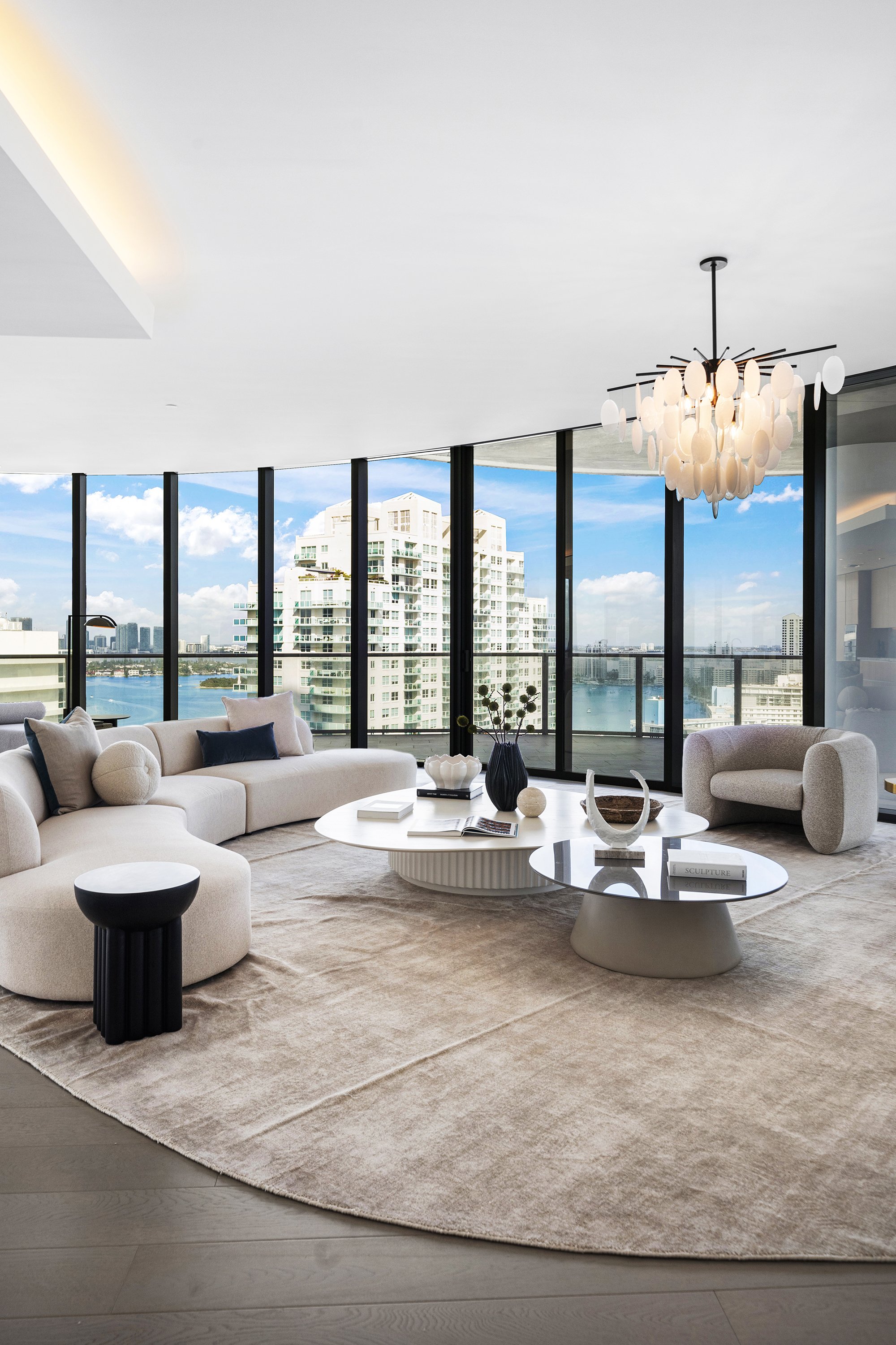 Modern living room with large windows, curved beige sofa, round coffee tables, neutral decor, contemporary chandelier, and city skyline view.