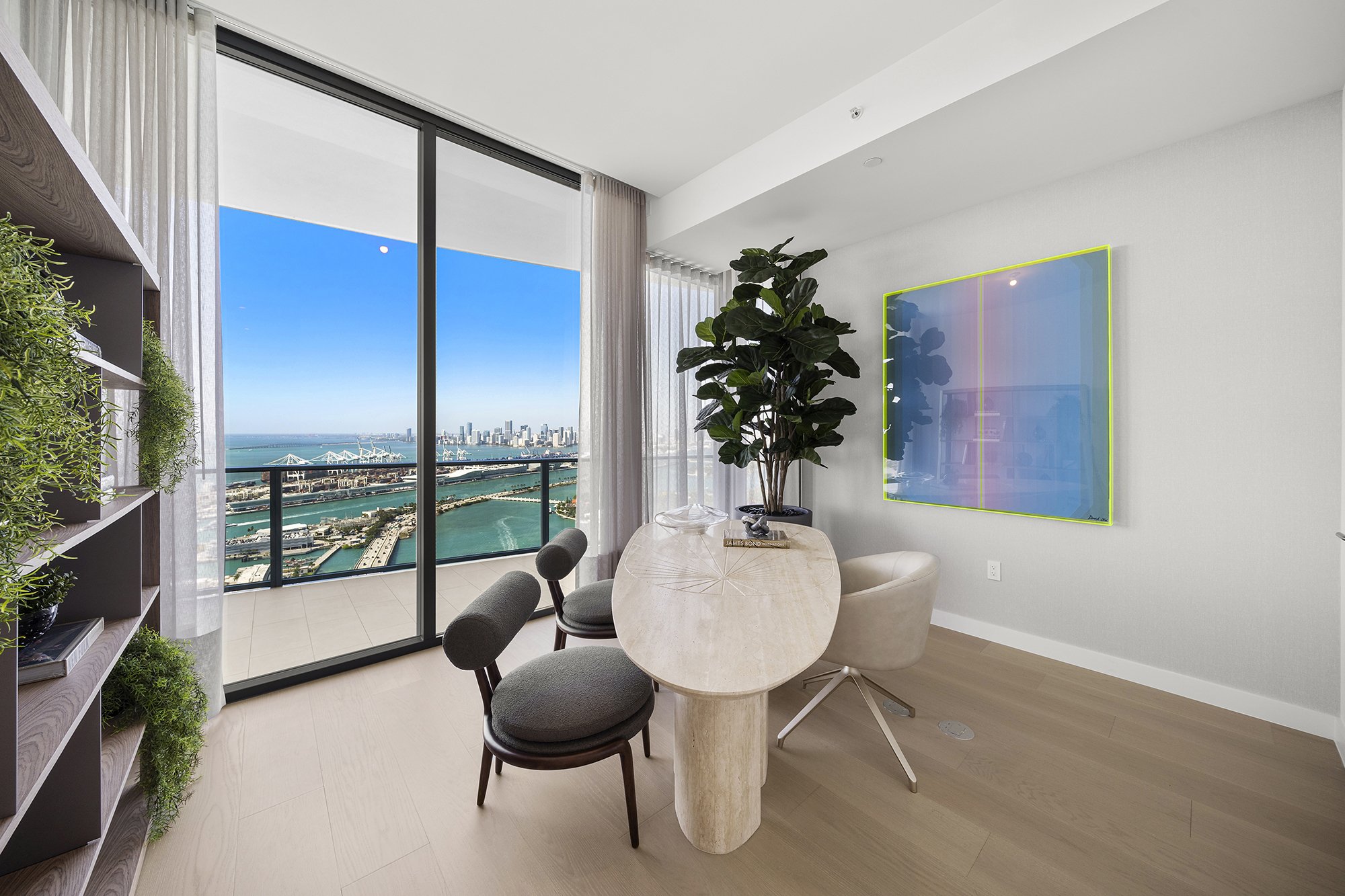 Modern living room with elegant furniture, large windows showing city and ocean view, indoor plants, and contemporary artwork on the wall.