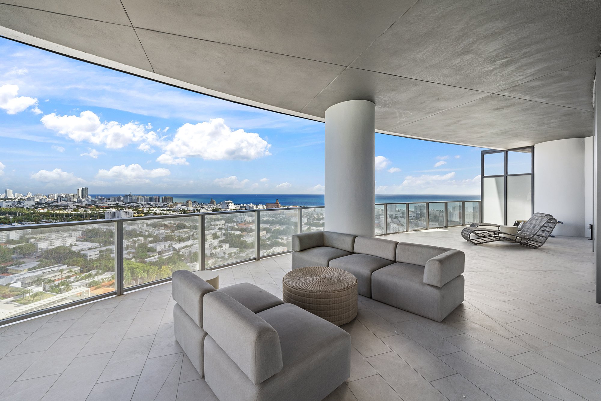 Modern rooftop terrace with gray seating area and round wicker table, overlooking cityscape and ocean view.
