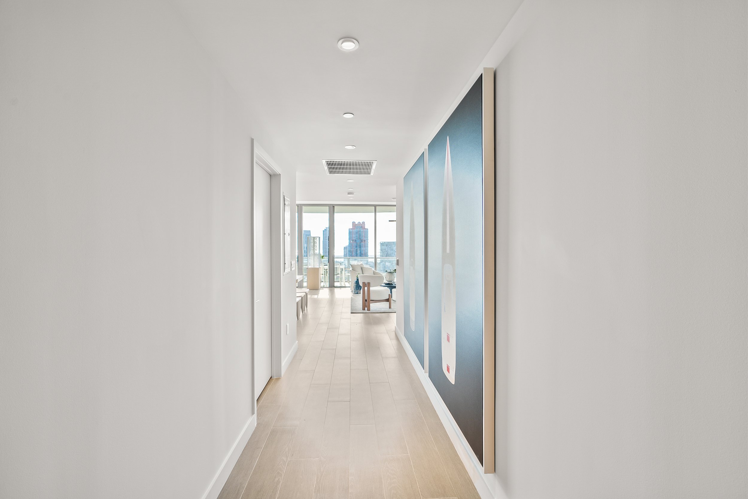 Modern apartment hallway with wooden flooring, abstract artwork on walls, and view of city skyline through large windows at the end.