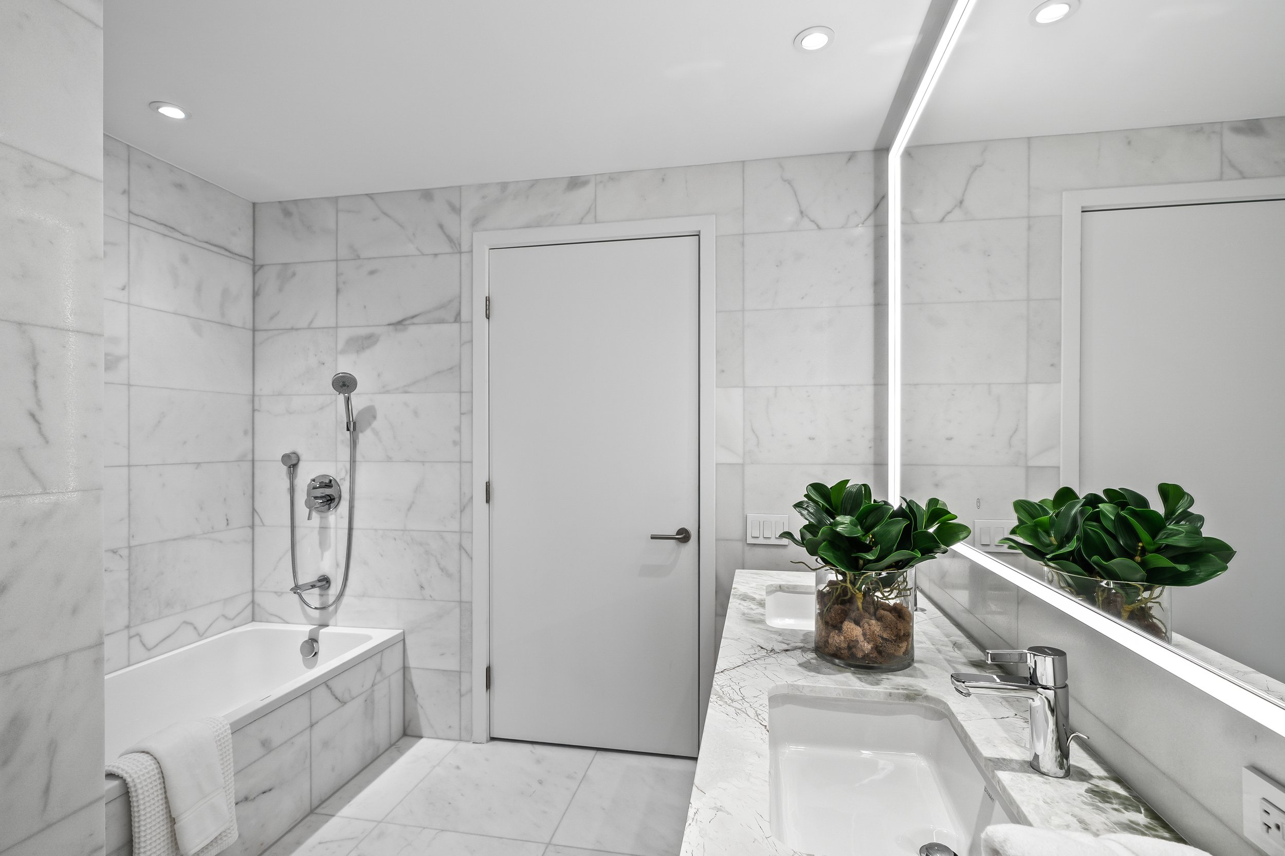 Modern bathroom with marble tiles, a bathtub, white door, vanity with sink, and a potted plant on the counter.