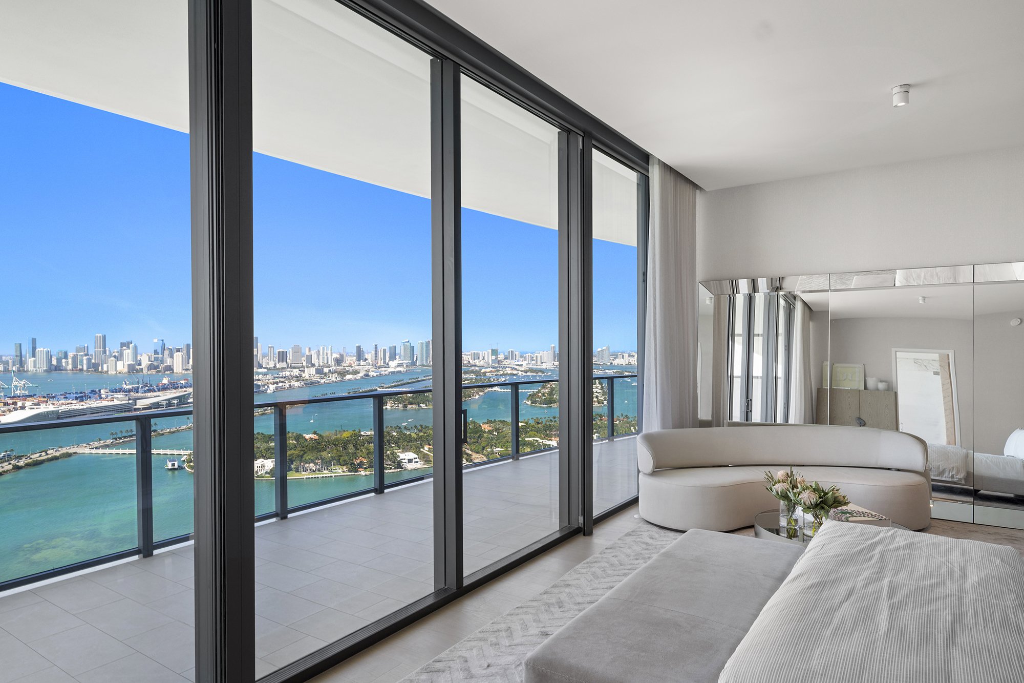 Modern luxury living room with expansive floor-to-ceiling windows offering a panoramic view of a city skyline and waterway, featuring sleek furniture and minimalist decor.