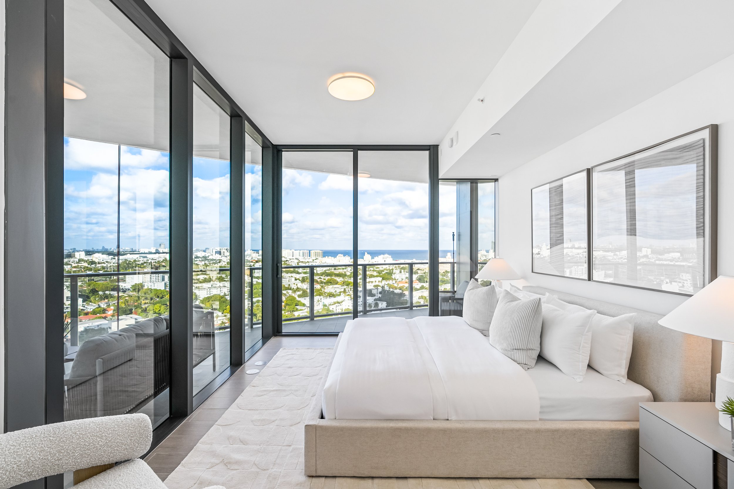 Modern bedroom with large windows offering a city and ocean view, featuring a bed with white linens, decorative pillows, and bedside lamps, minimalistic decor with framed art on the walls.