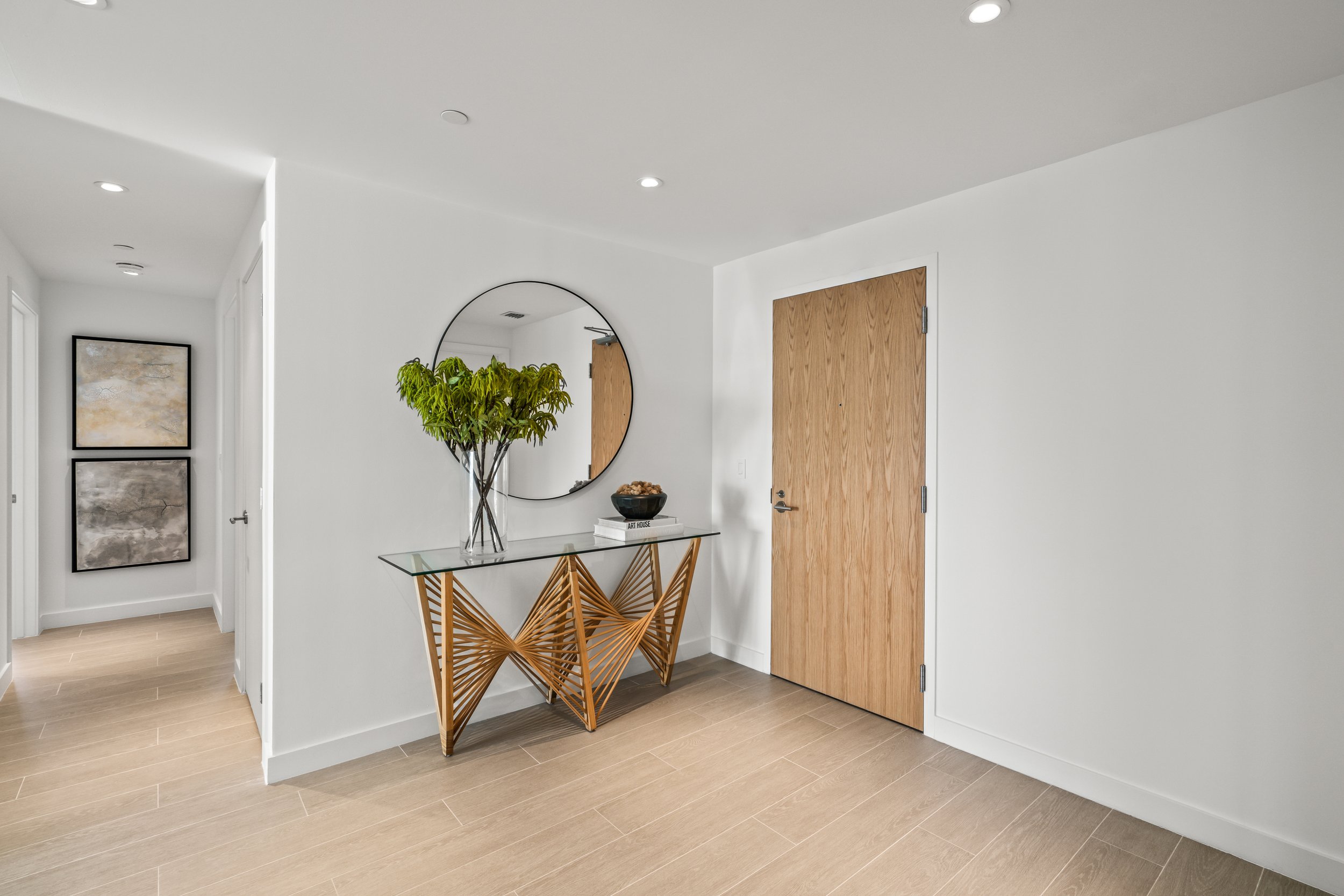 Modern hallway with wooden door, glass console table with decorative base, large round mirror, potted plant, and wall art.
