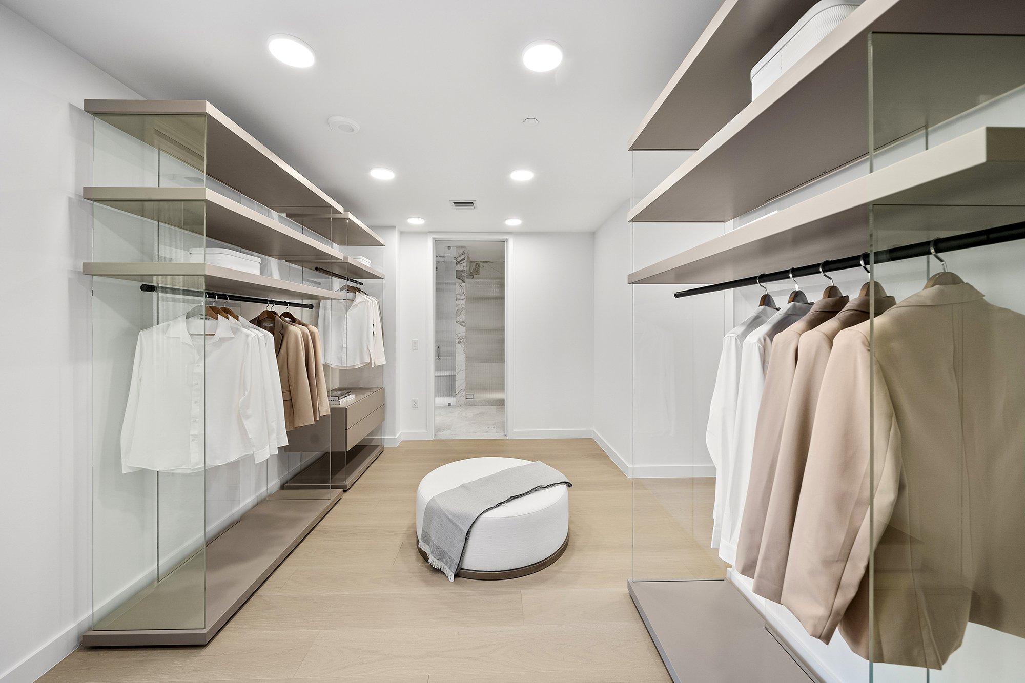 Modern walk-in closet with glass shelves, hanging clothes, and a round ottoman in the center, featuring neutral colors.