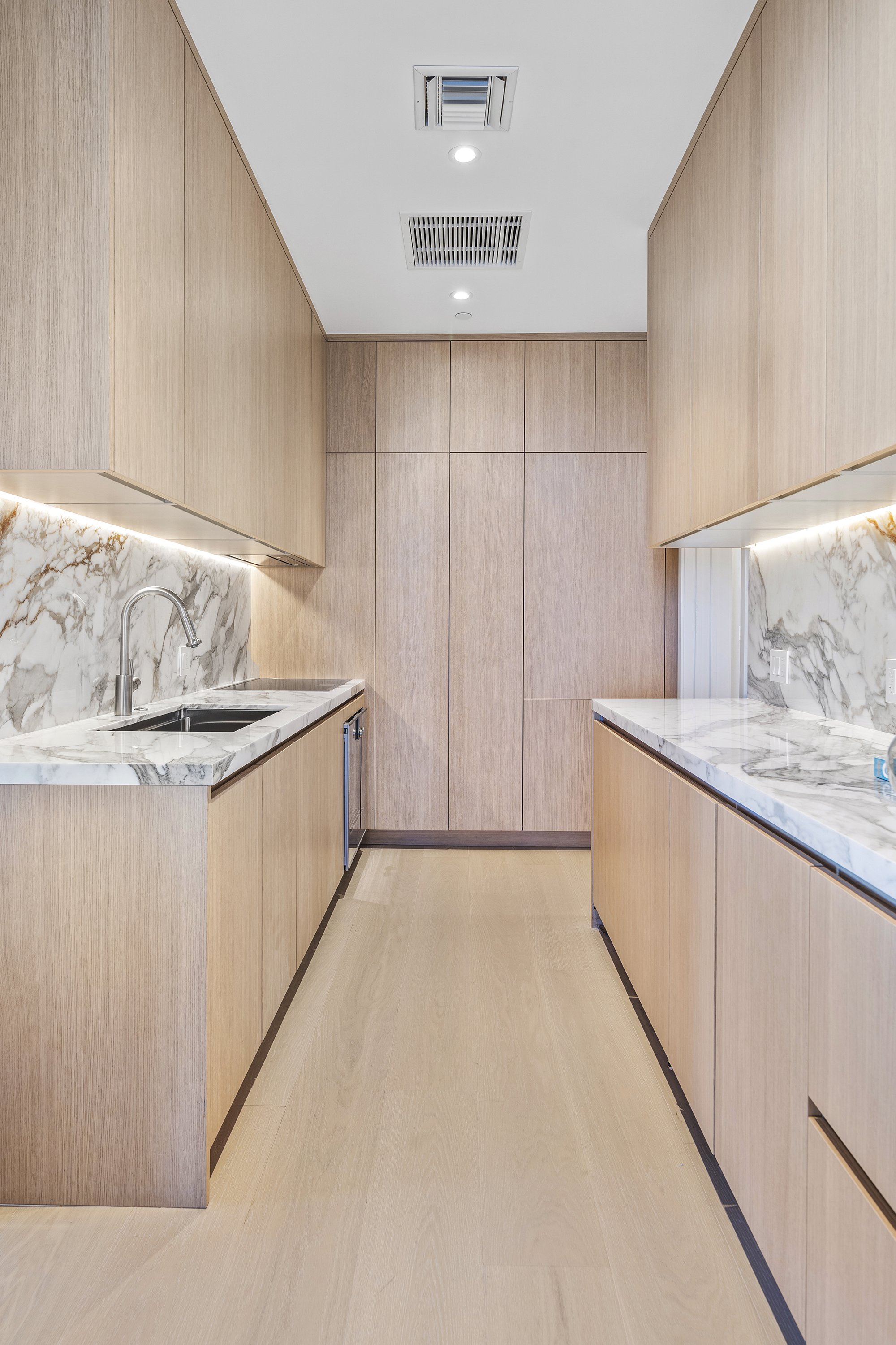 Modern kitchen featuring light wood cabinetry, marble countertops, and a marble backsplash. A built-in oven and sink are visible, with overhead lights illuminating the space.
