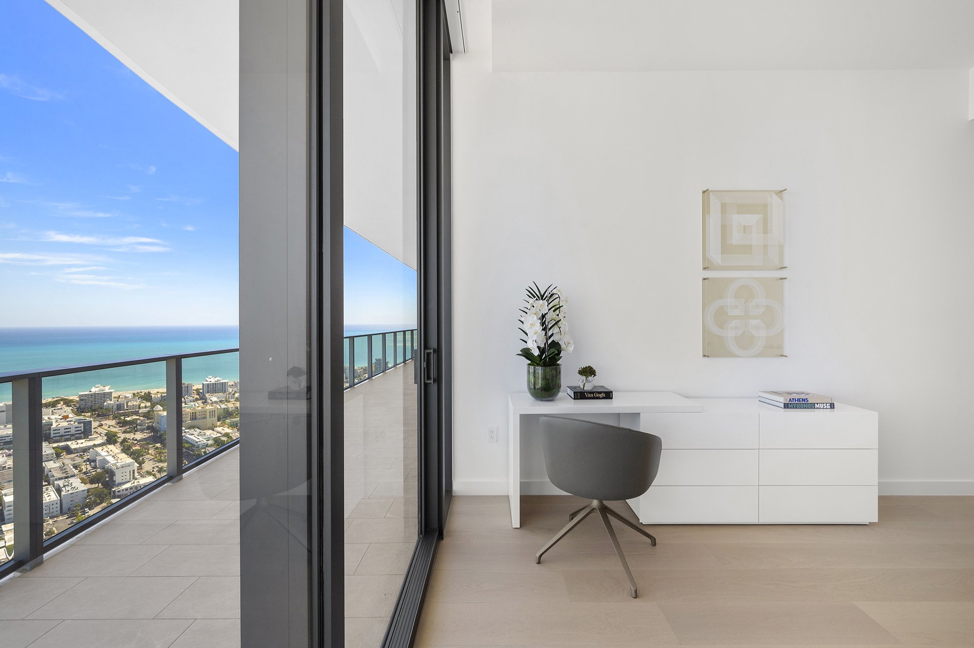 Modern home office with desk and chair next to a large window overlooking a cityscape and ocean view. The room has white walls, framed artwork, and decorative plants on the desk.