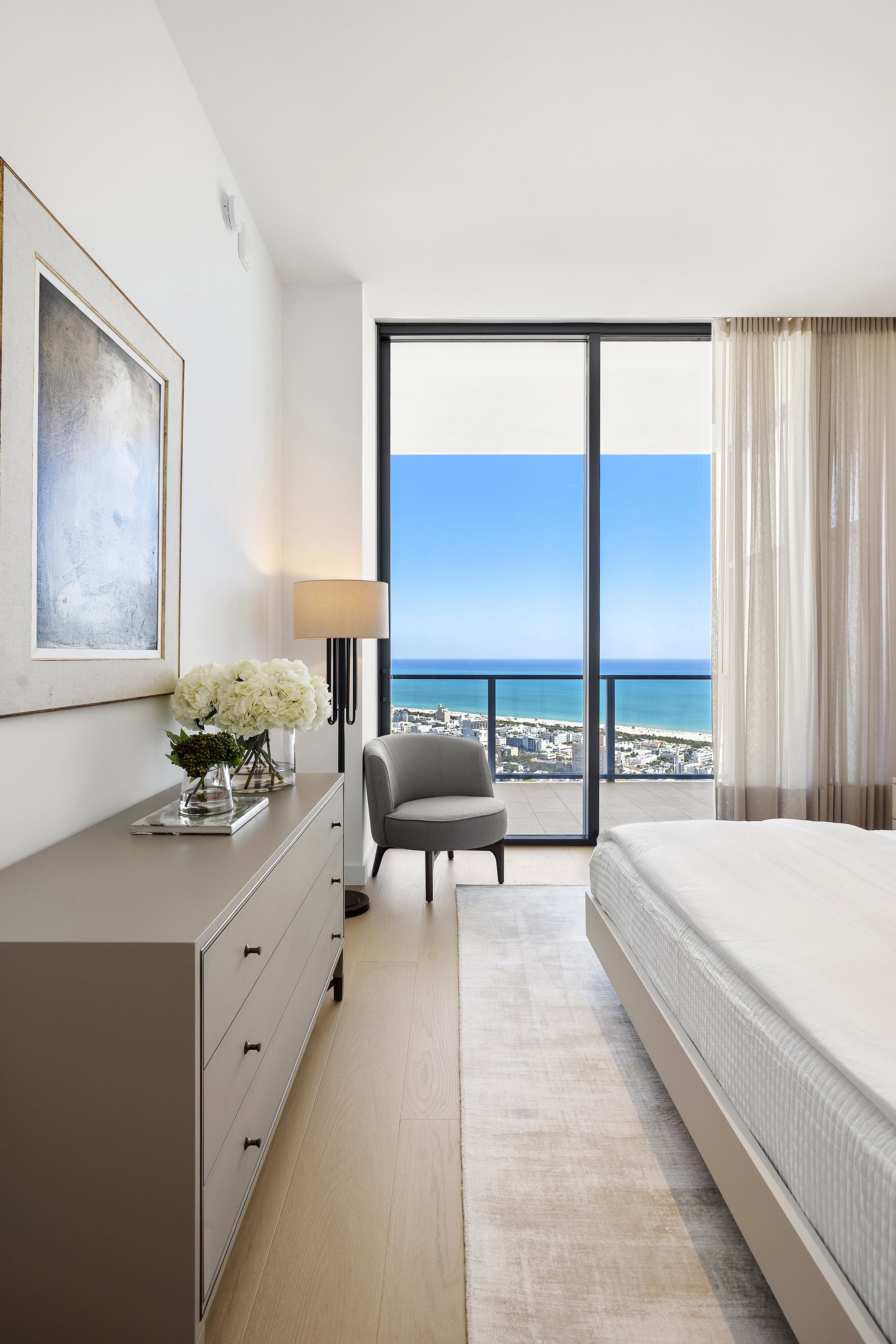 Modern bedroom with a dresser, a chair, a lamp, and a large window with ocean view.
