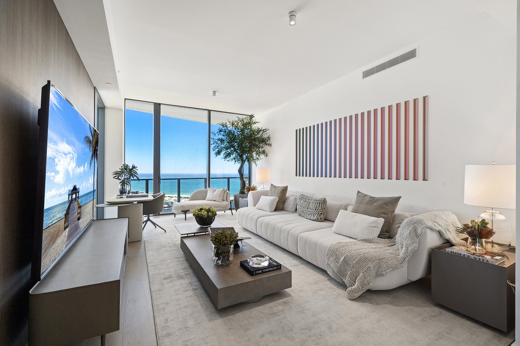 Modern living room with beige sofa, coffee table, large TV, and ocean view through floor-to-ceiling windows.