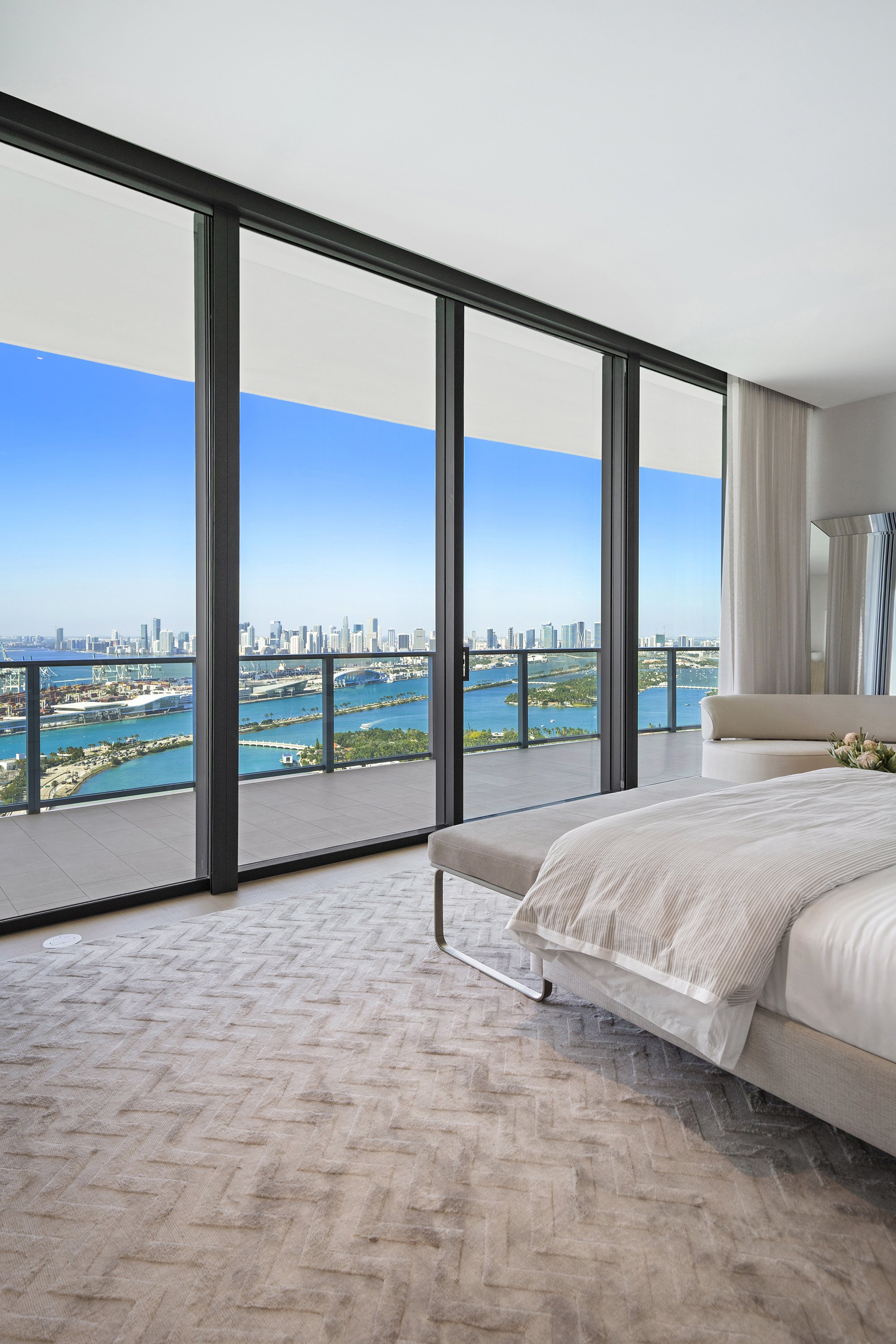 Modern bedroom with large windows, ocean view, city skyline, and stylish furniture.