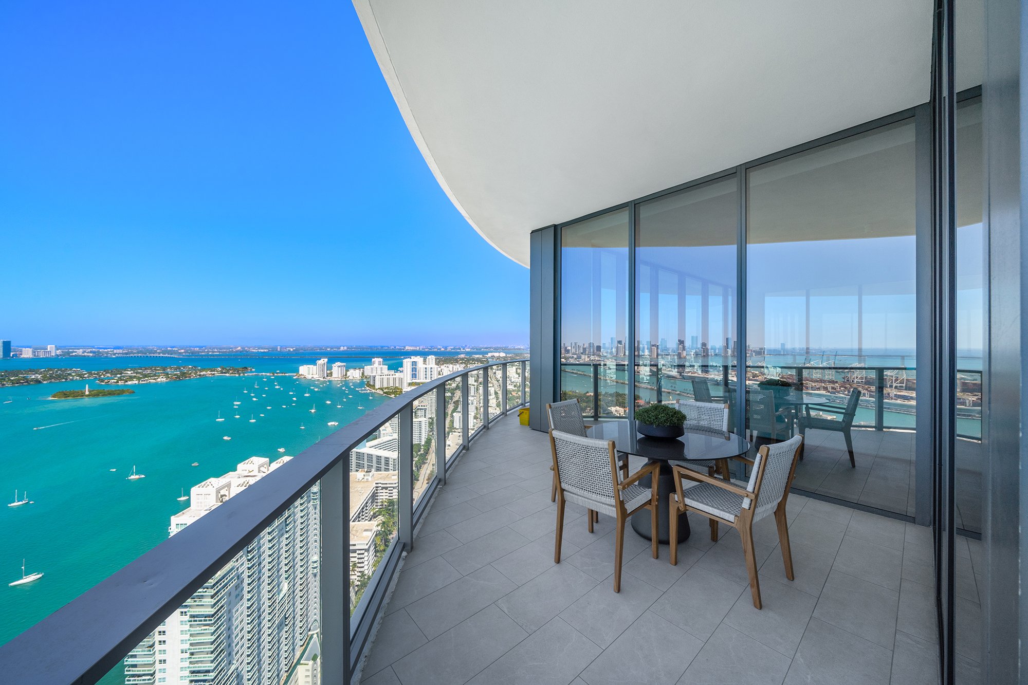 High-rise balcony overlooking blue ocean and cityscape with patio furniture.
