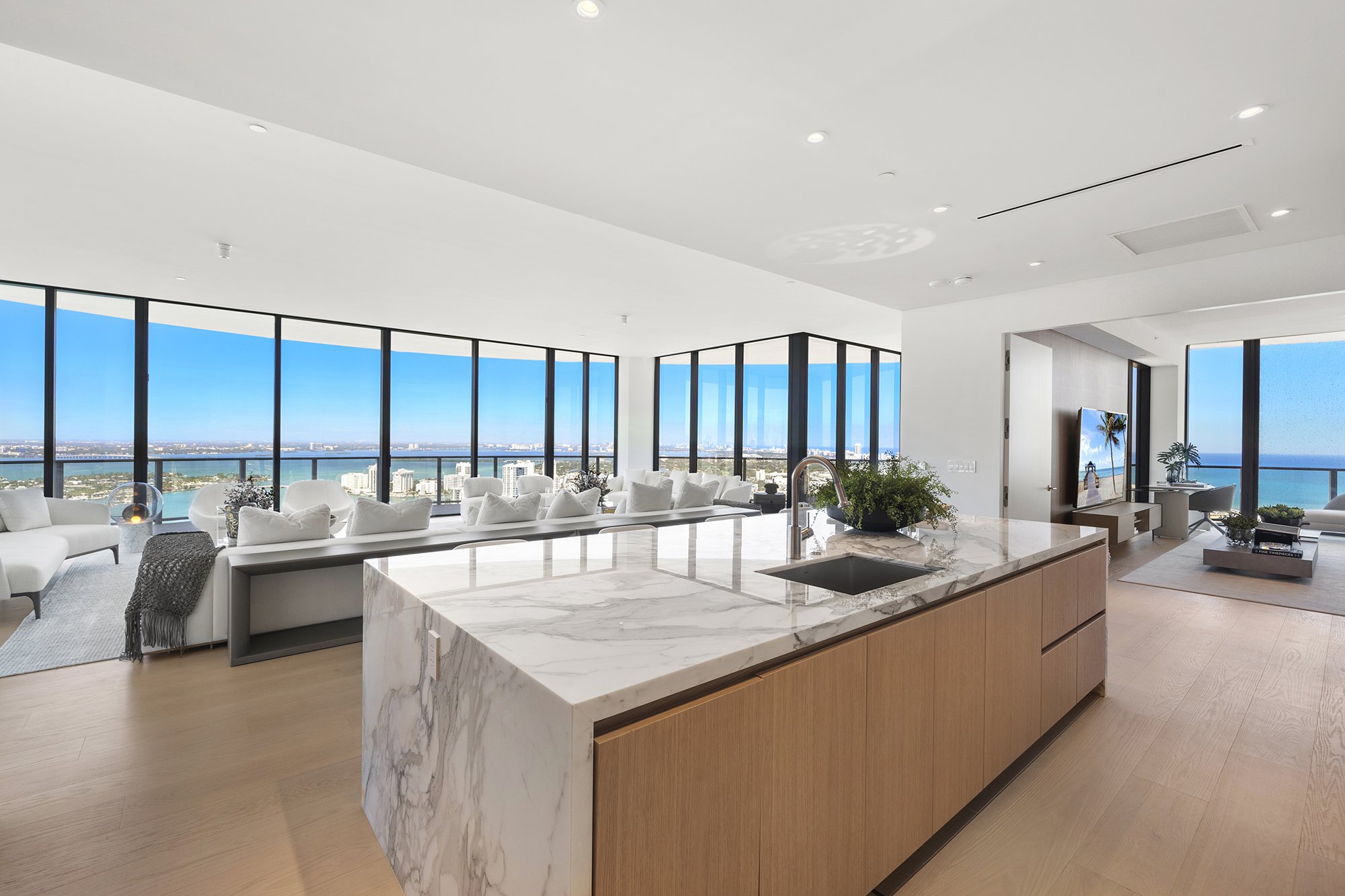 Modern apartment interior with large windows offering a cityscape and waterfront view, featuring a marble kitchen island, wooden cabinets, white couches, and a TV area.