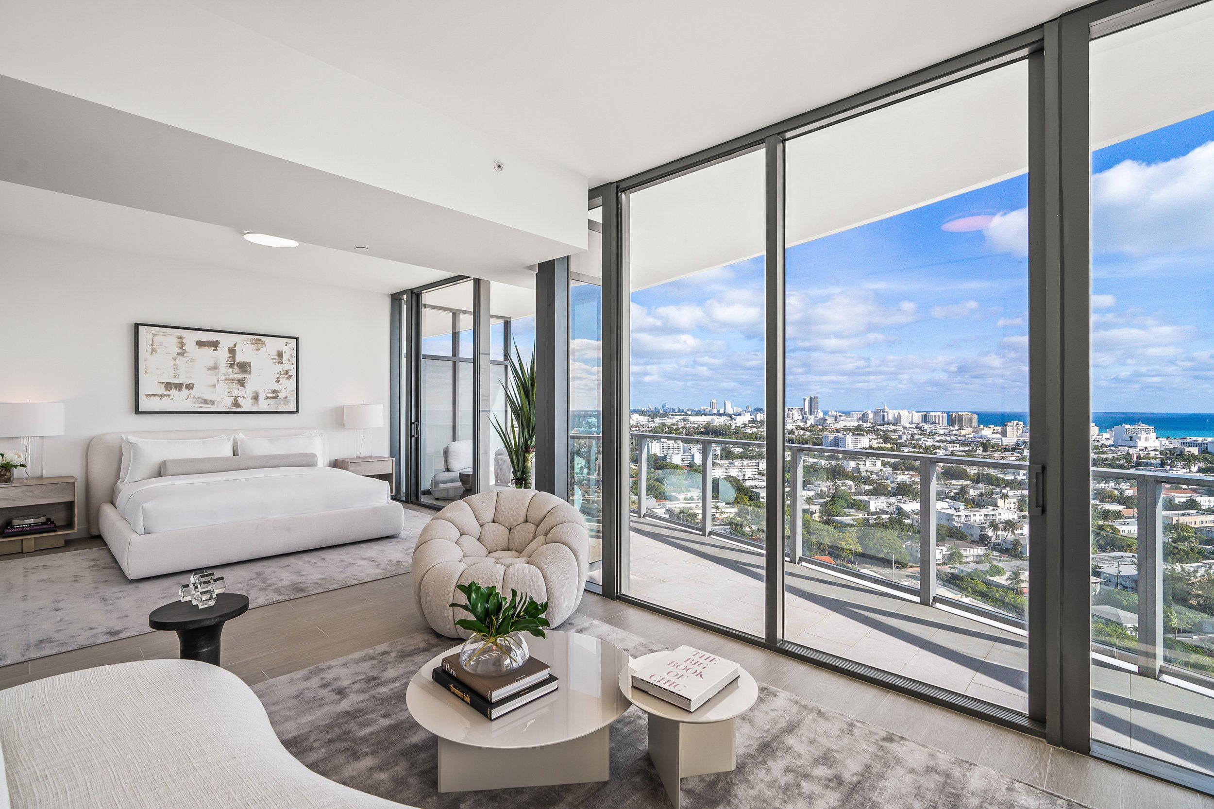 Modern minimalist bedroom with large windows, a white bed, neutral furniture, and a city view.