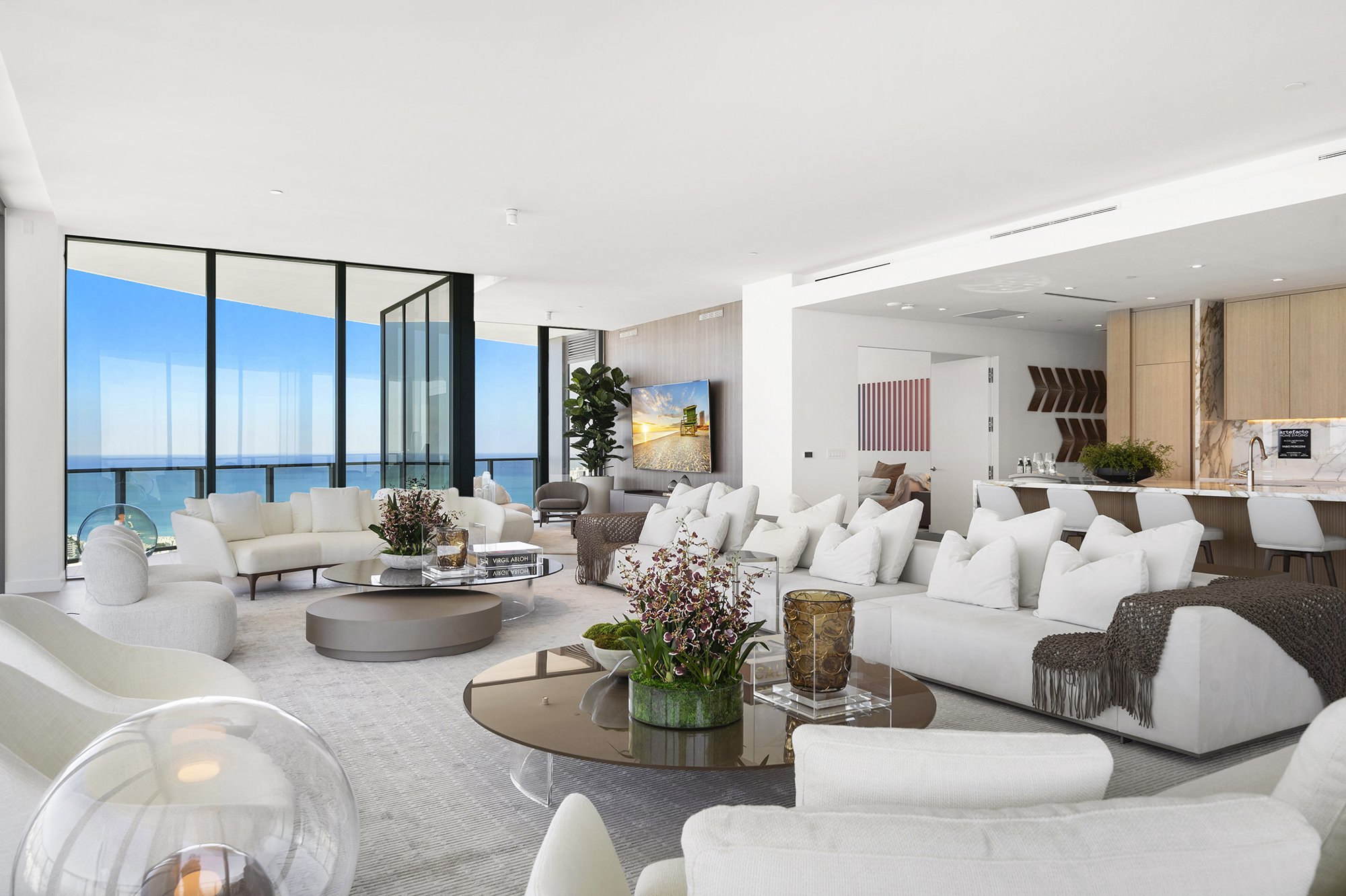 Luxury modern living room with ocean view, featuring white sofas, glass coffee tables with plants, large windows, open kitchen with bar stools, and decorative elements.