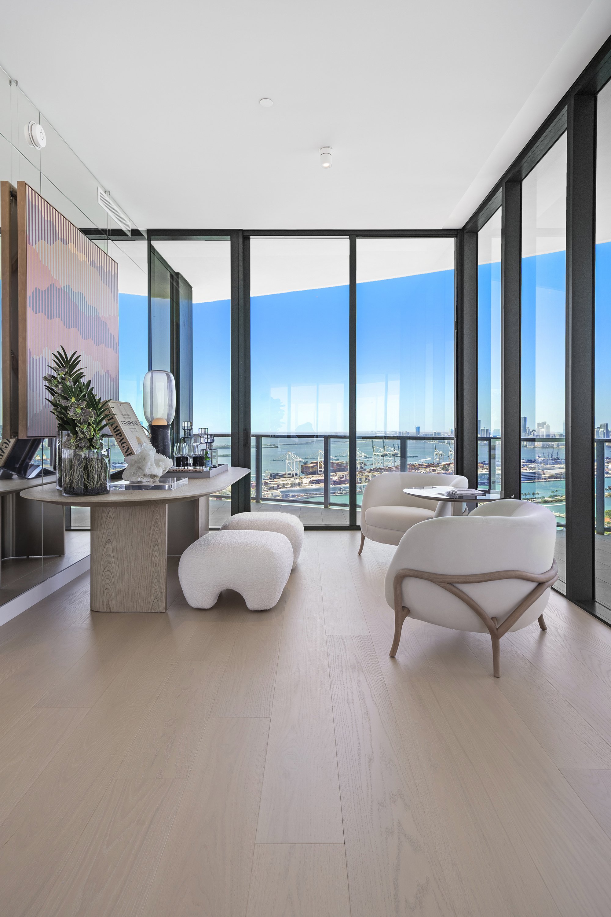 Modern living room with floor-to-ceiling windows, light wood flooring, contemporary chairs, a small round table, decorative plants, and artwork. The view outside shows a cityscape and ocean.