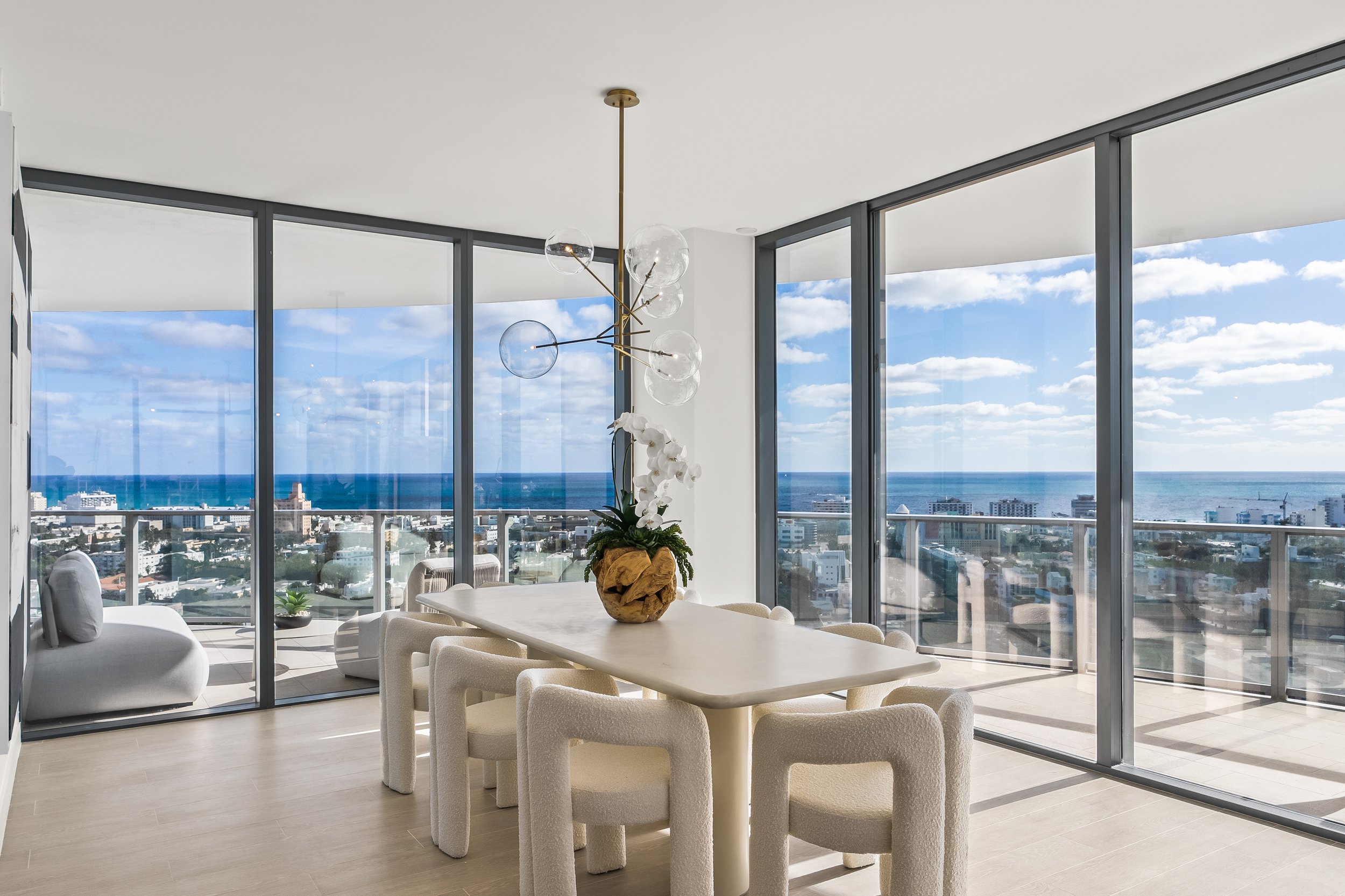 Modern dining room with glass walls offering ocean view, featuring a long table surrounded by chairs, a potted plant centerpiece, and a contemporary chandelier.