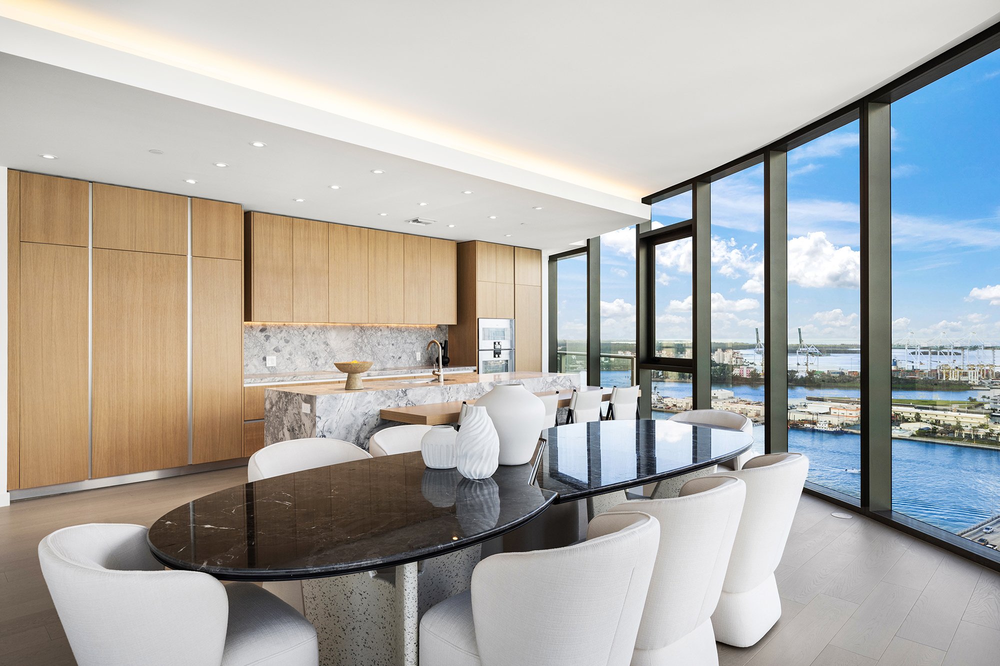 Modern kitchen and dining area with large windows offering a waterfront view, featuring wooden cabinetry, a marble island, a black dining table with white chairs, and decorative vases.
