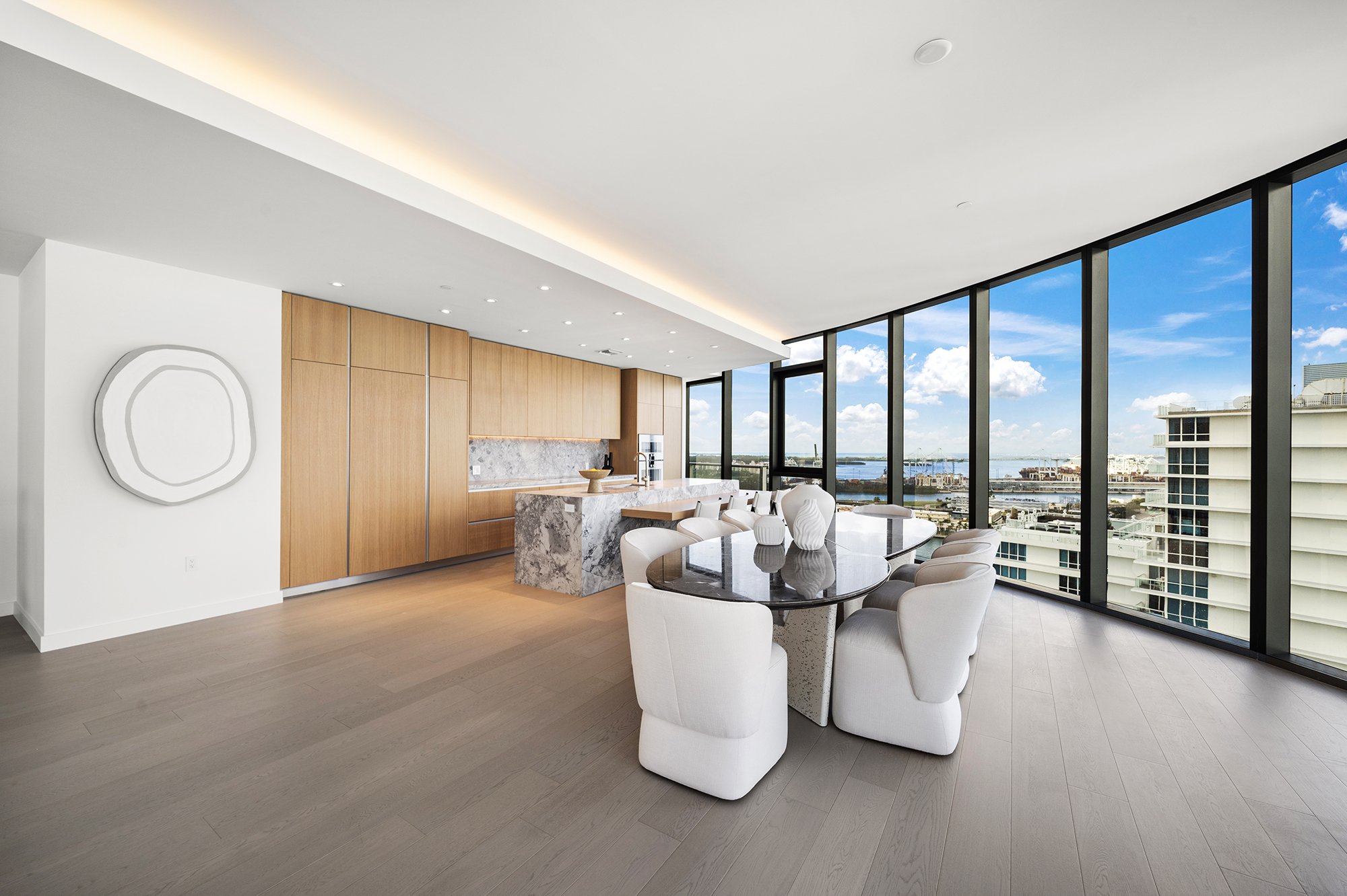 Modern luxury apartment interior with panoramic windows, wooden cabinetry, a marble kitchen island, and a dining area with white chairs and a glass table.