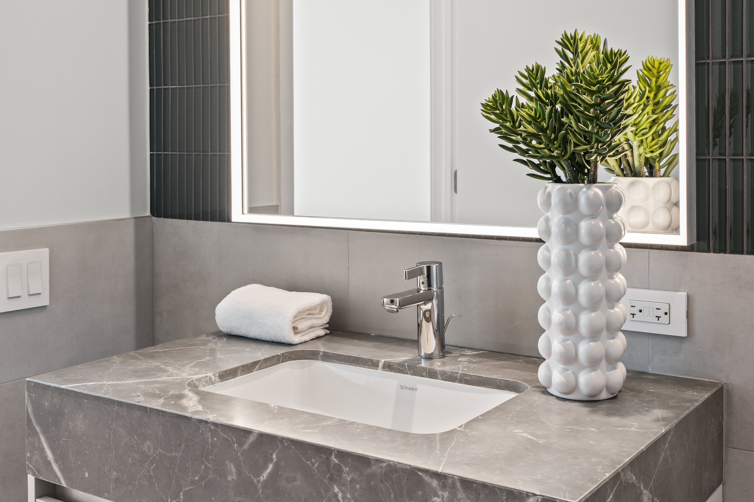 Modern bathroom sink with gray marble countertop, a white decorative vase with green plants, a chrome faucet, and a folded white towel near a backlit mirror.
